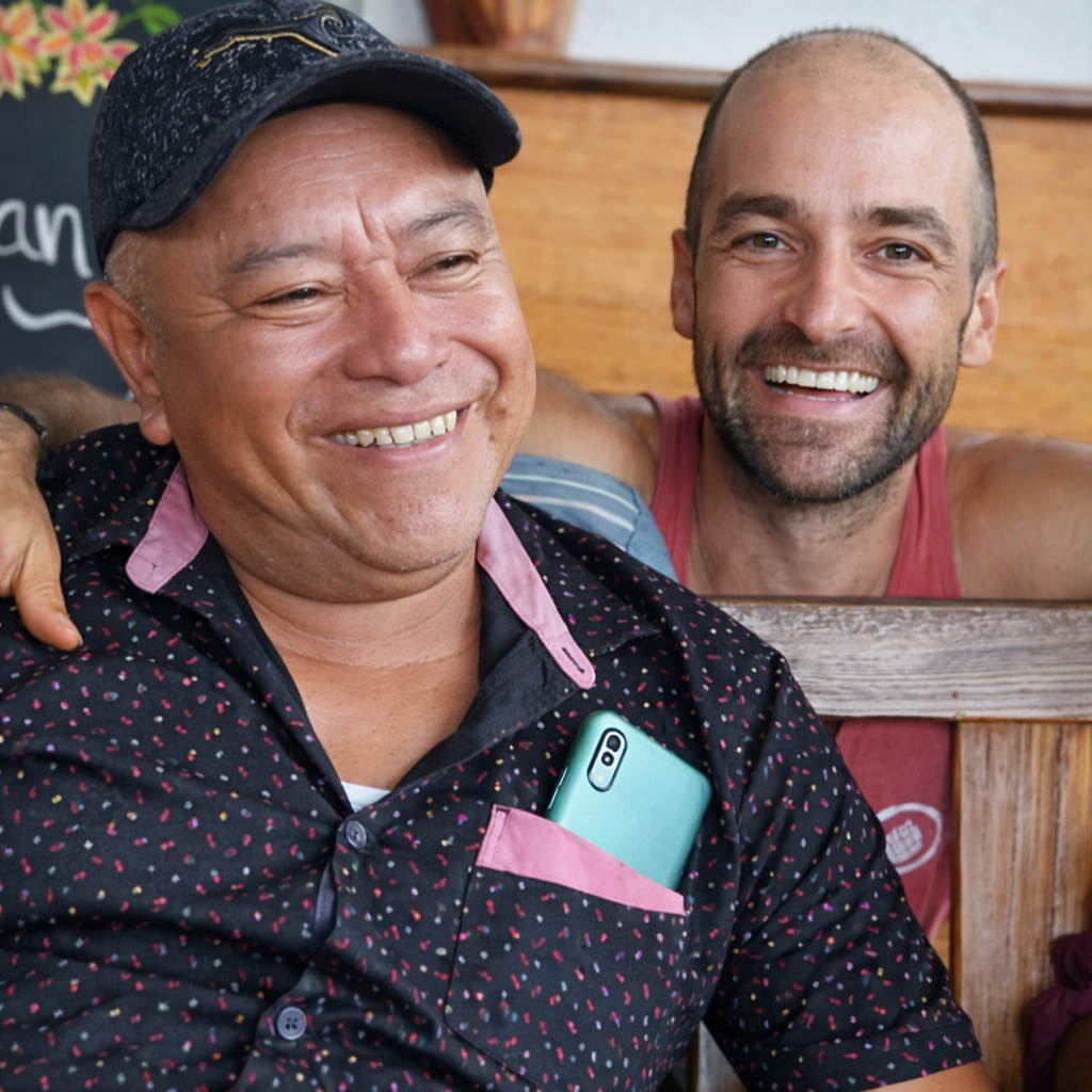 Two men smiling and sitting close together in a casual indoor setting, one wearing a black cap and the other without hair, both appearing happy.