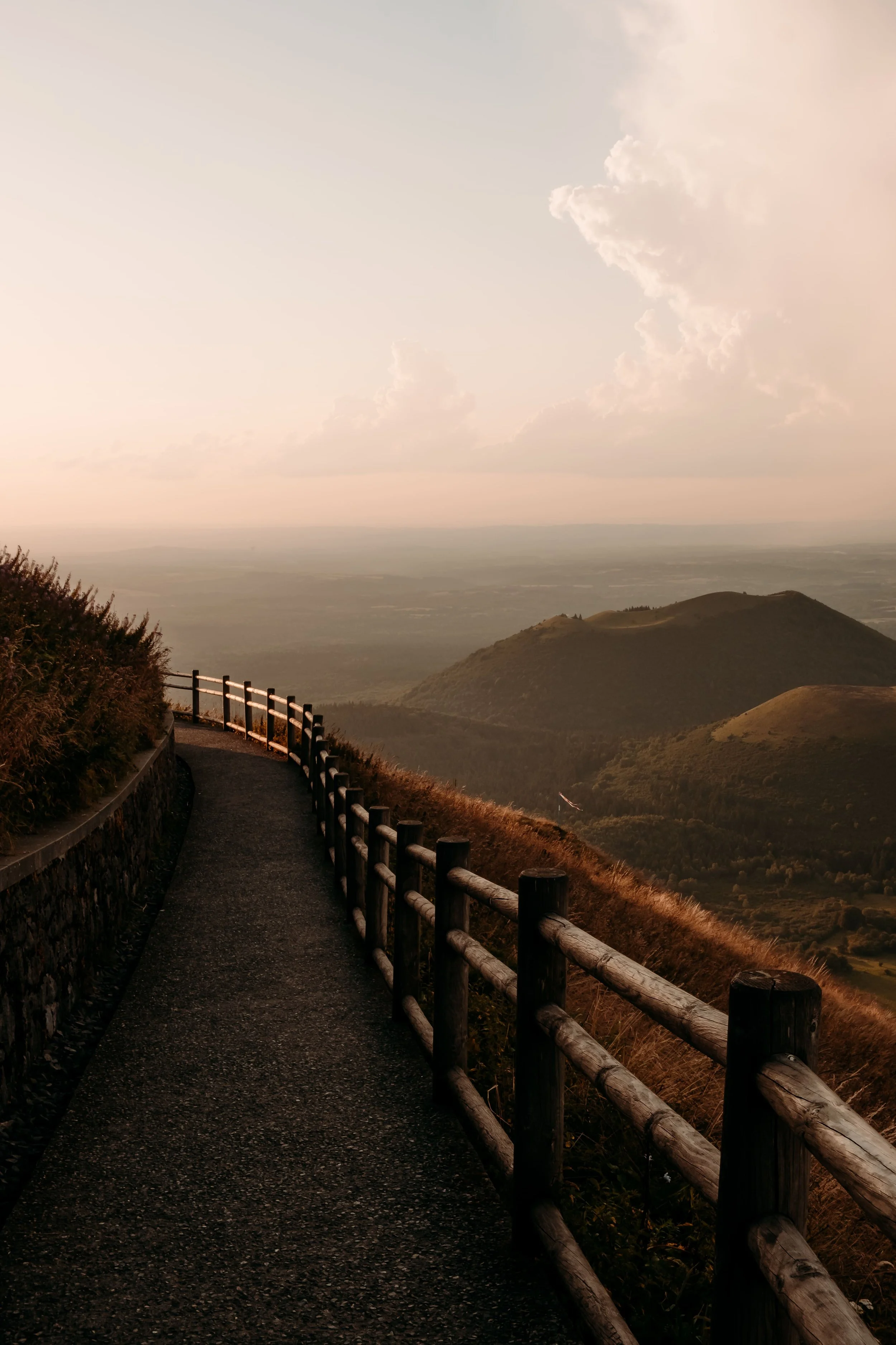 Chemin de montagne serpentant avec une clôture en bois, vue sur des collines et un ciel partiellement nuageux au crépuscule.