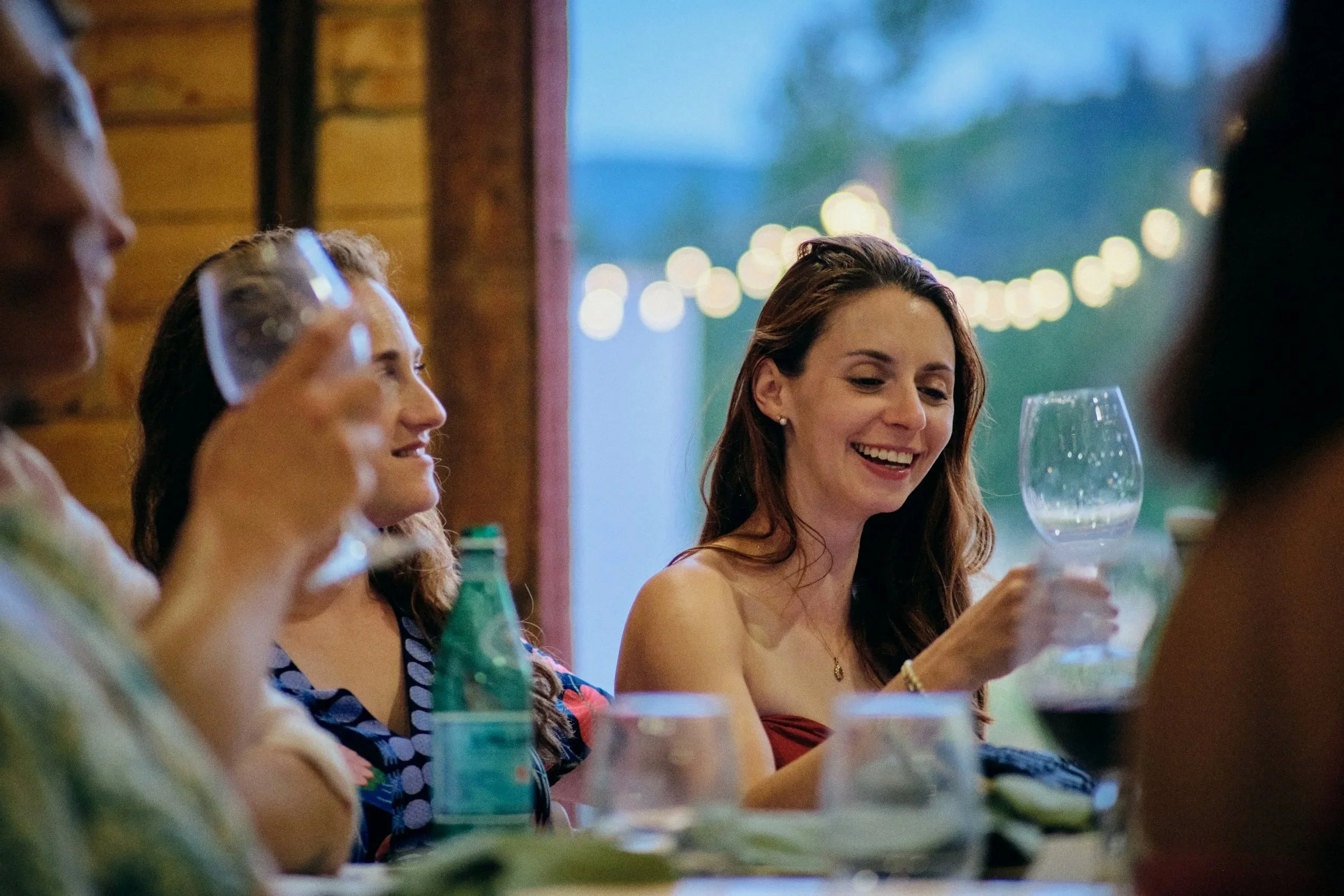 Guests laughing over wine at a winery dinner under string lights