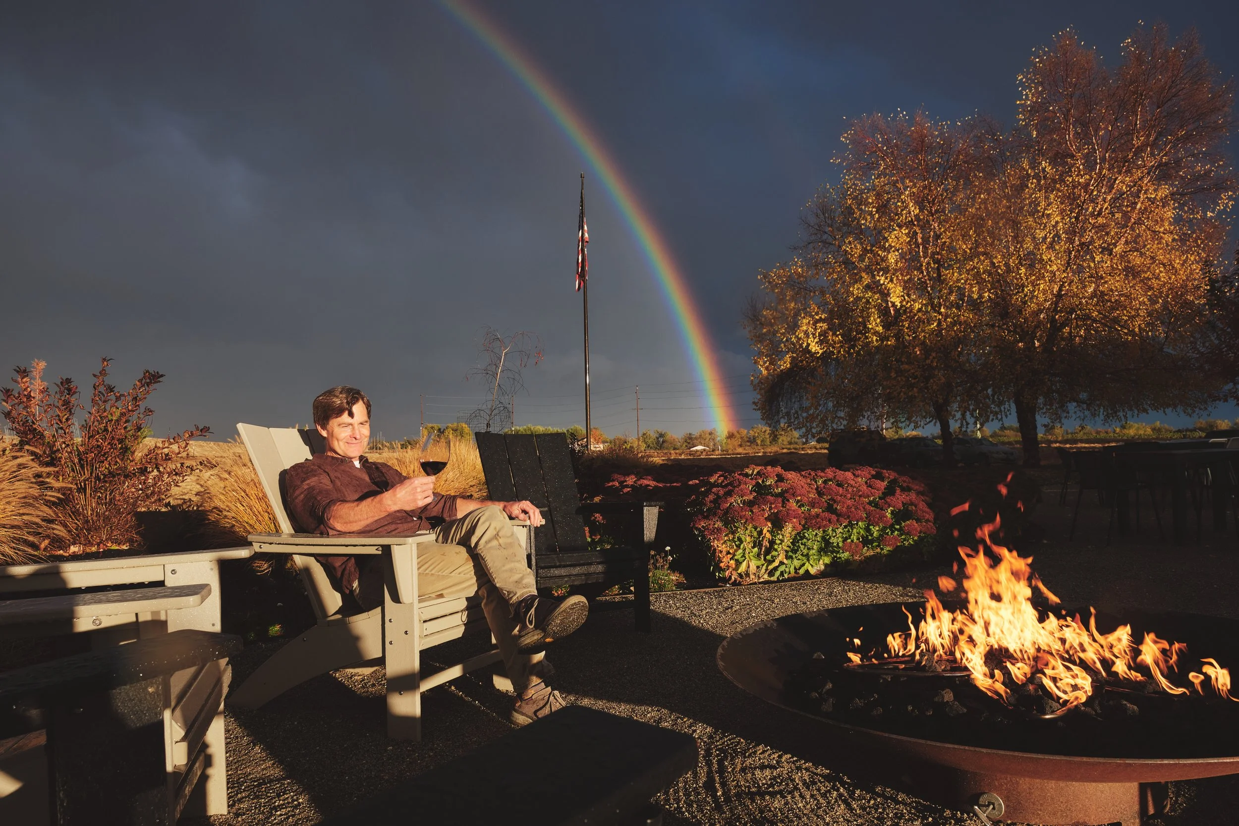 Man relaxing with a glass of wine by a fire pit under a rainbow at a winery