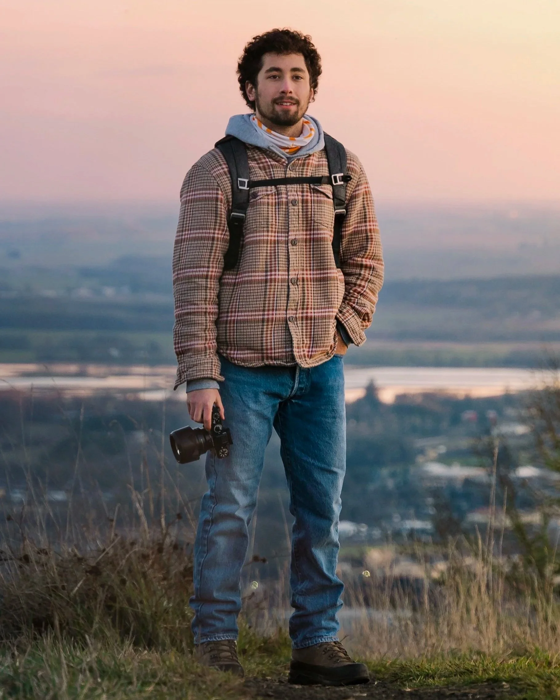 A young man standing outdoors on a hill at sunset, wearing a plaid jacket, gray hoodie, and carrying a backpack, holding a camera in his right hand with a city view in the background.