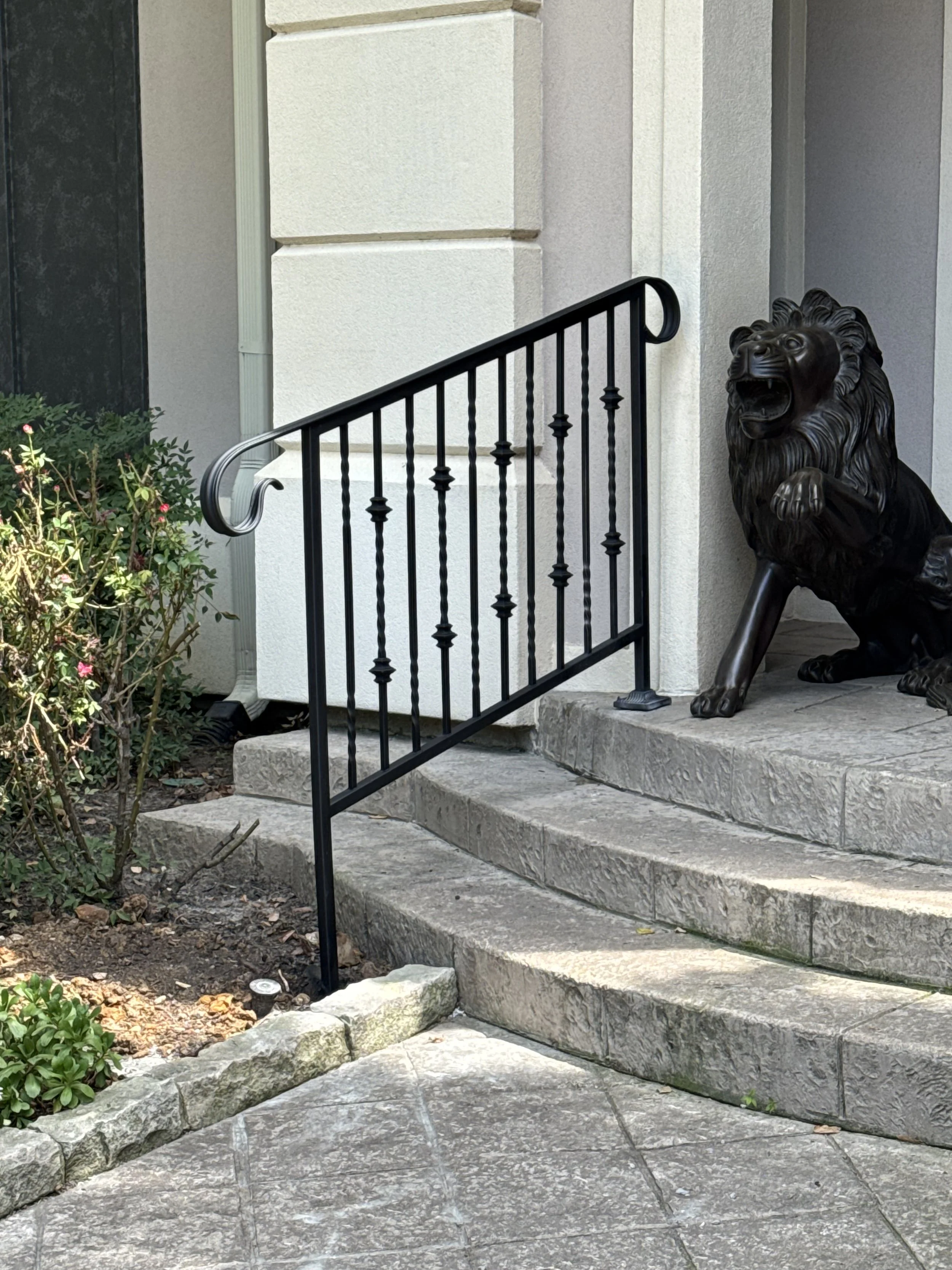 A stone staircase leading to a building entrance with a metal railing on one side. A bronze lion statue is positioned at the bottom of the stairs, facing outward with an open mouth. There are some bushes with pink flowers on the left side of the stai