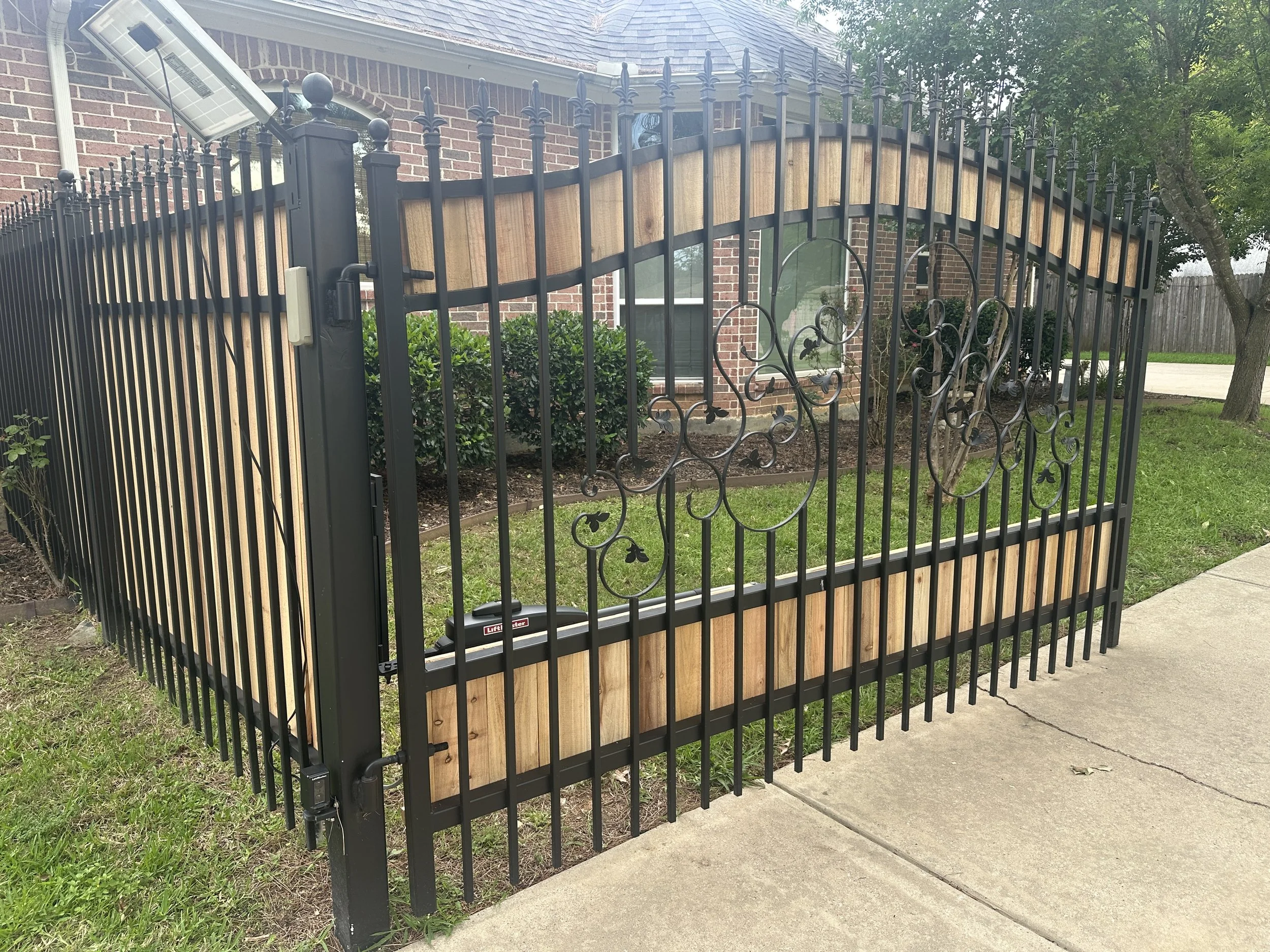 A black metal fence with decorative ironwork surrounds a house, with a sun panel attached to the fence. The house has brick walls, arched windows, and a shingled roof. There are green bushes and trees nearby, and a sidewalk and grass in the foregroun