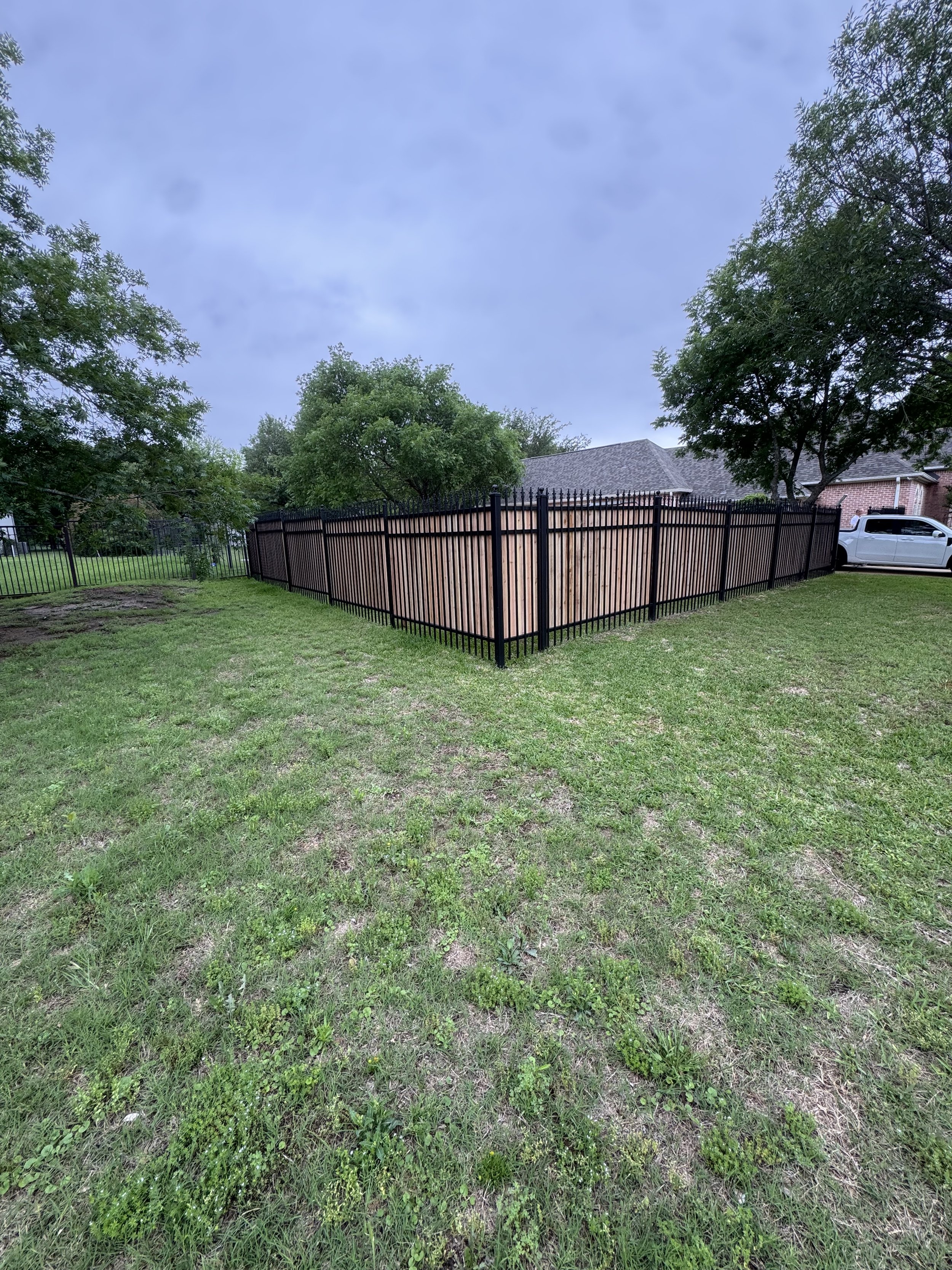 A fenced backyard with a newly installed wooden fence with black metal posts, surrounded by green grass and trees, with a gray house and a silver truck in the background under a cloudy sky.