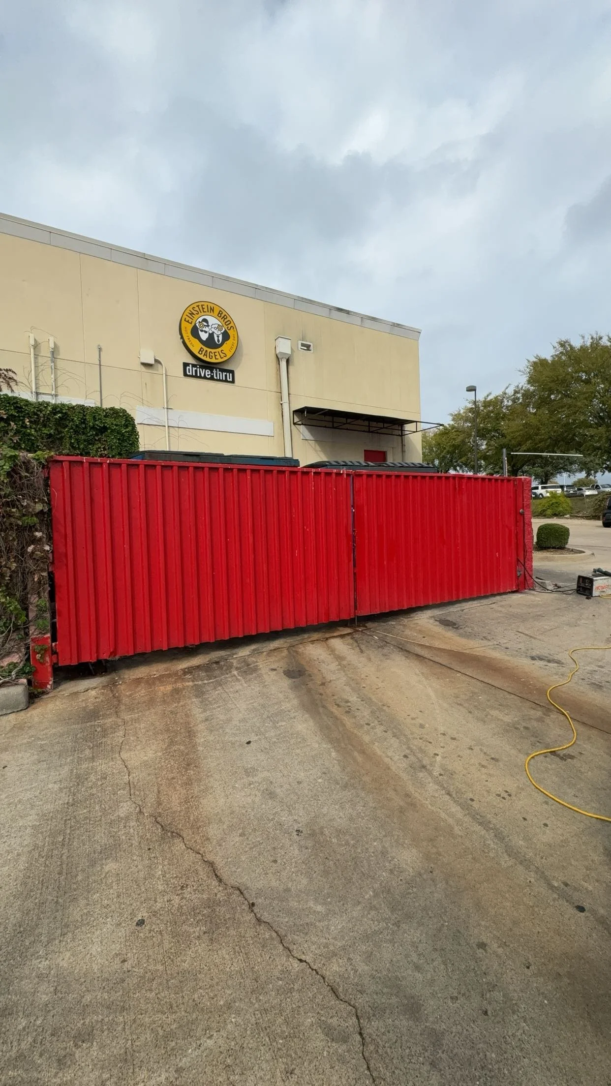 . In front of the building, there is a red metal fence, and the parking lot appears to be under construction or maintenance. The sky is cloudy, and there are some trees and parking spaces in the background.