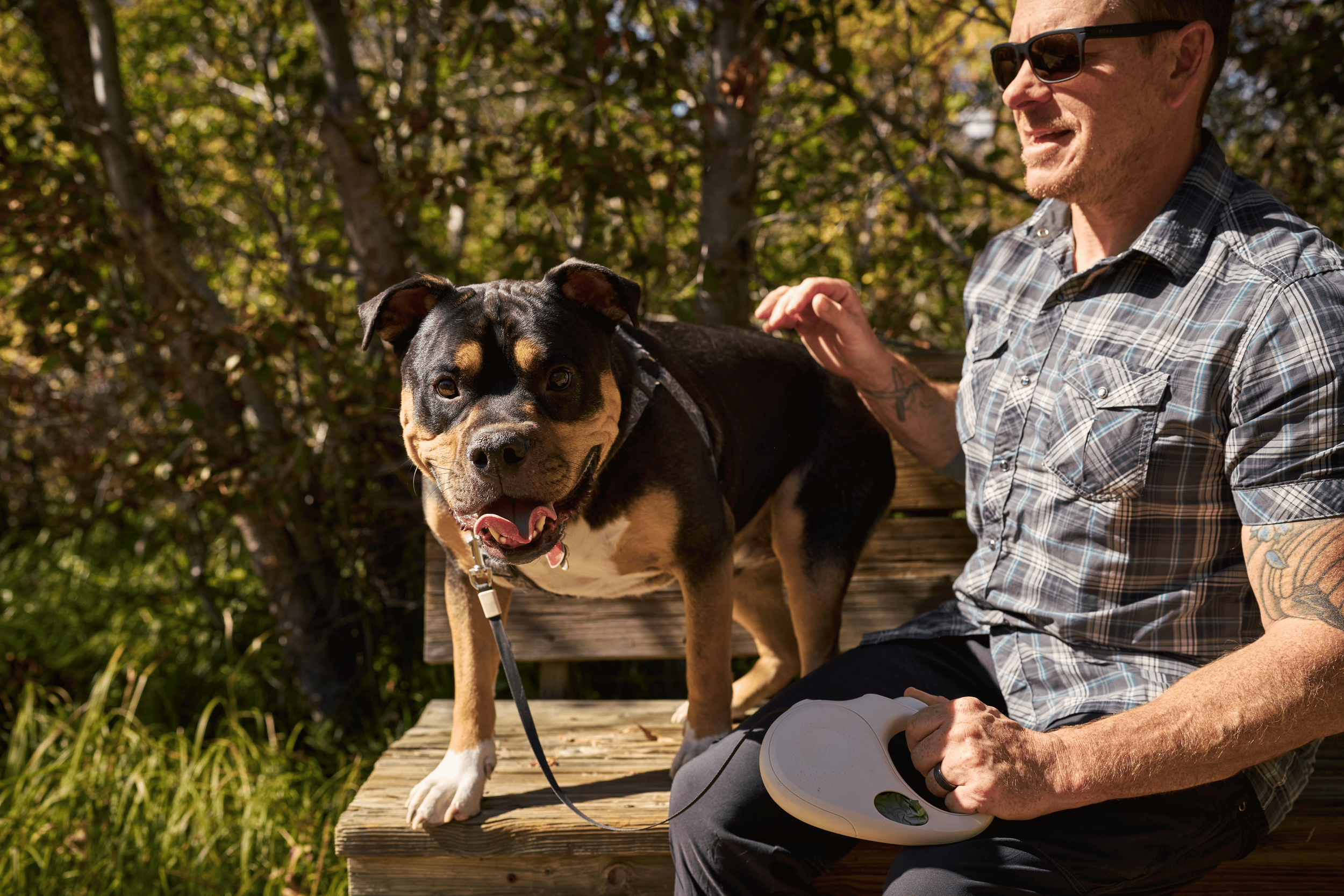 A man wearing sunglasses and a plaid shirt sitting on a wooden bench outdoors, petting a happy black and tan dog on a leash, with trees in the background - Dog Product Video Production