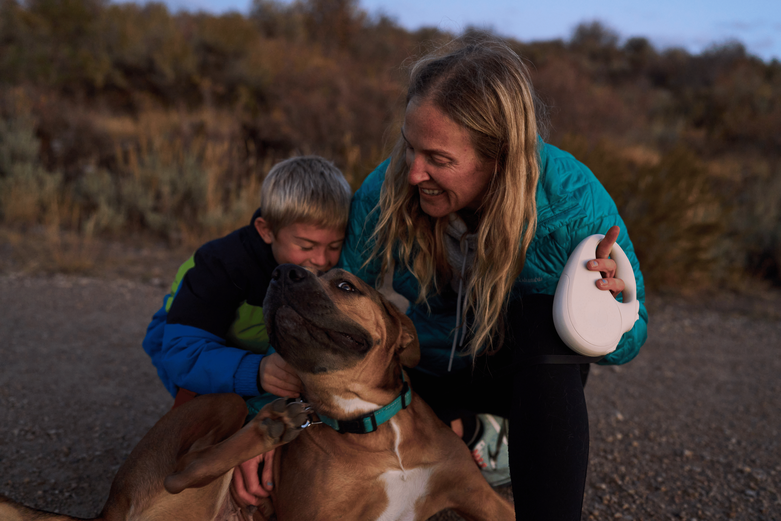 A woman and a young boy are hugging a large brown dog outdoors during dusk, with a background of trees and a cloudy sky- Dog Product Video Production