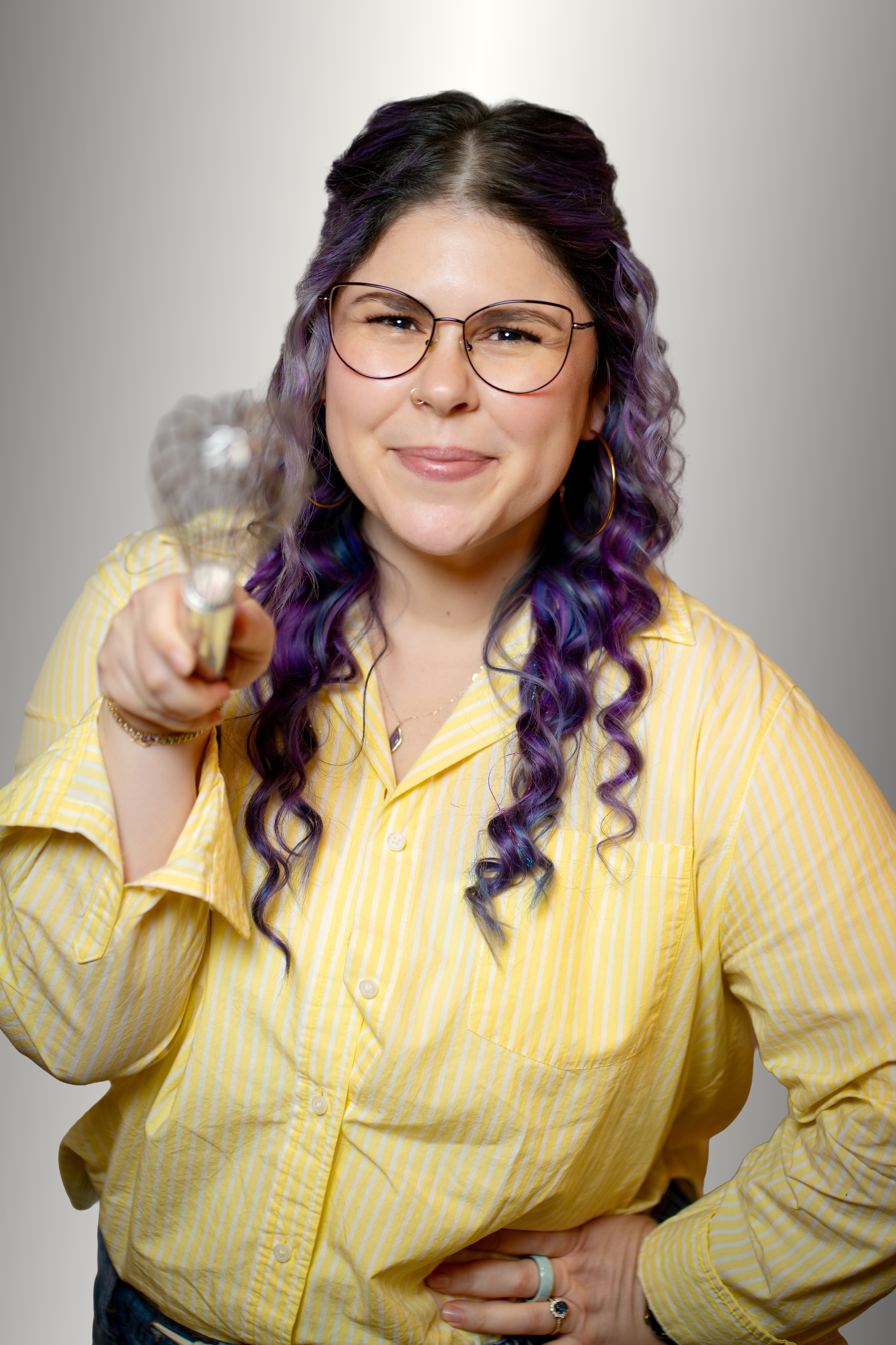 Woman with glasses and red hair sitting, wearing a floral dress and cream cardigan, against a backdrop of leafy artwork.