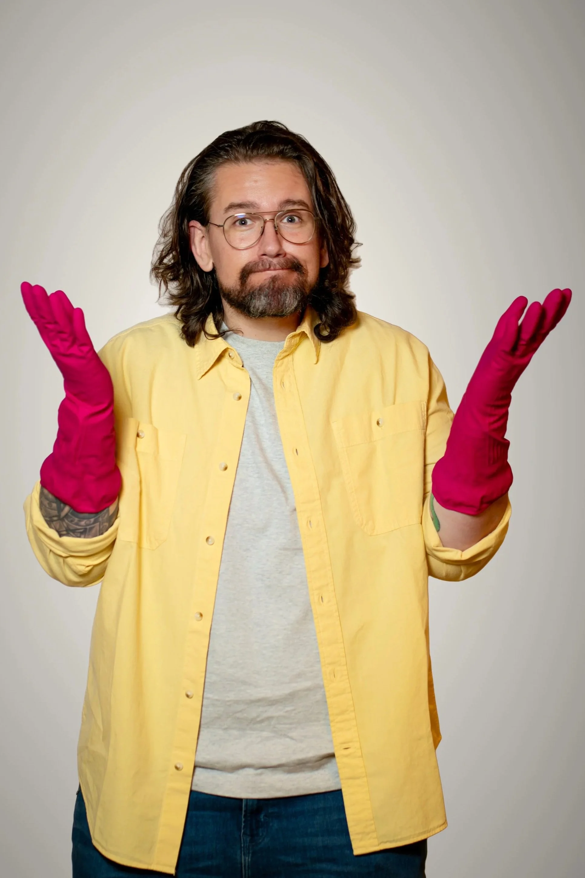 Man with long hair and glasses, wearing a black polo and smartwatch, standing with arms crossed in front of a wall with framed art.