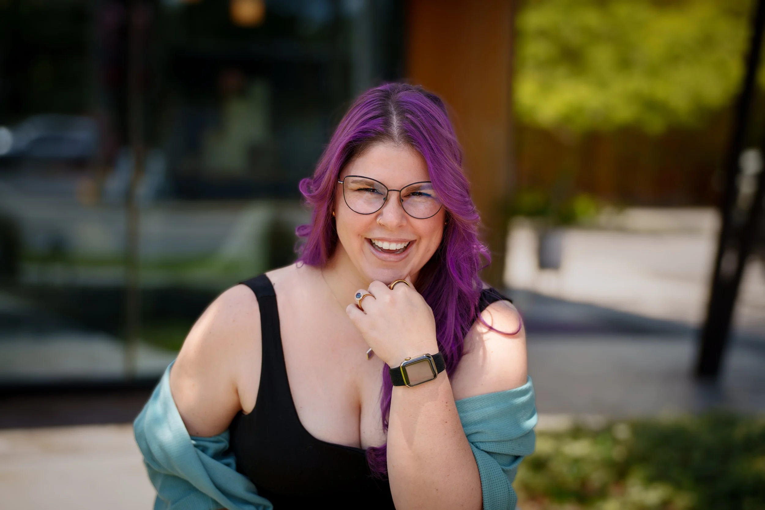 Woman with glasses and red hair sitting, wearing a floral dress and cream cardigan, against a backdrop of leafy artwork.