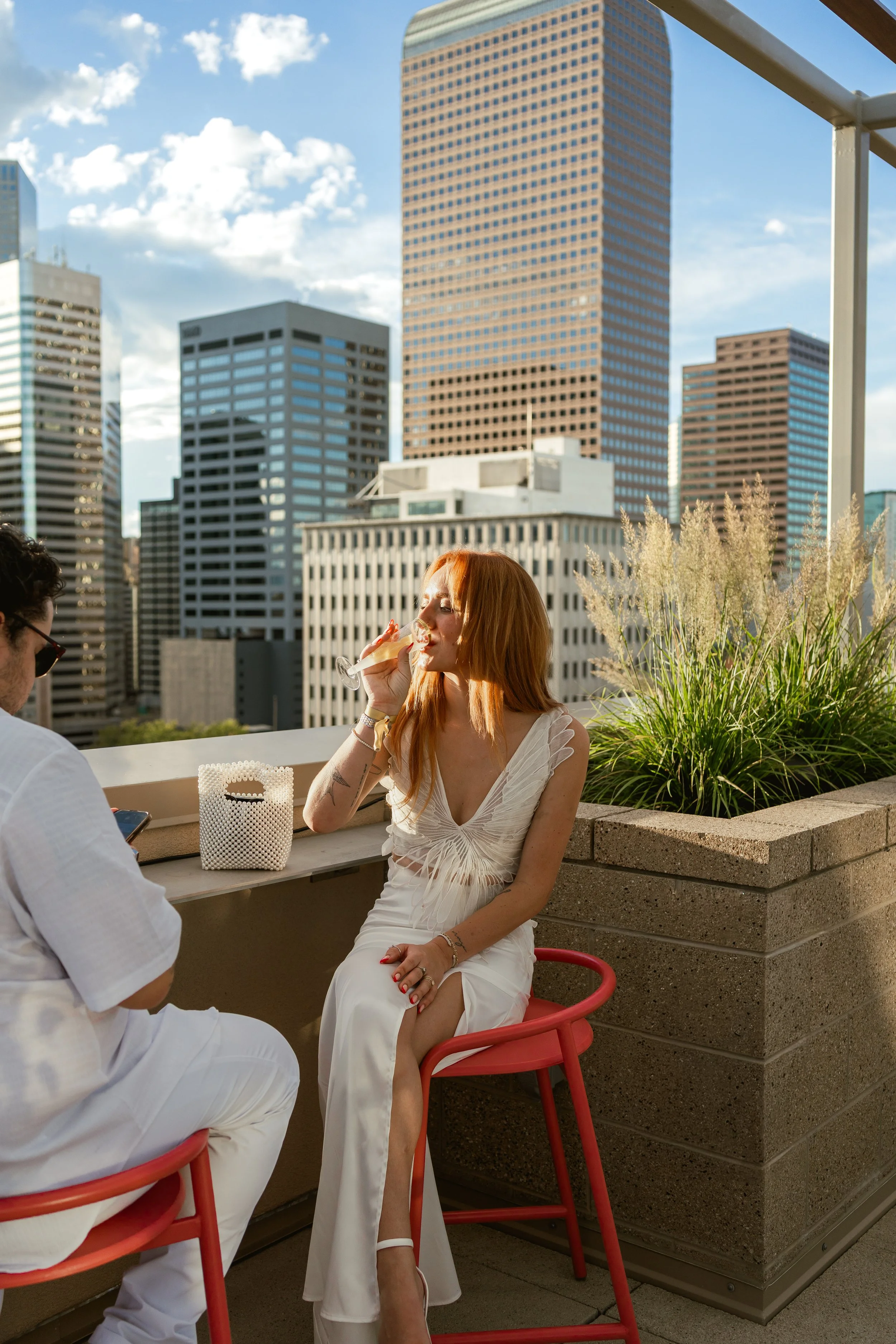 A woman with red hair wearing a white dress sitting on a red chair on a rooftop patio with a city skyline in the background, drinking from a glass. A man in a white shirt is sitting next to her, looking at his phone.