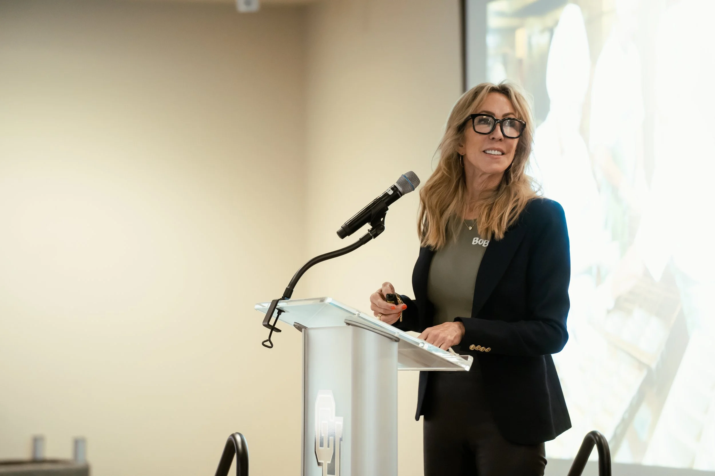 A woman with long blonde hair, glasses, and a gray shirt stands at a podium with a microphone, smiling during a presentation or speech.