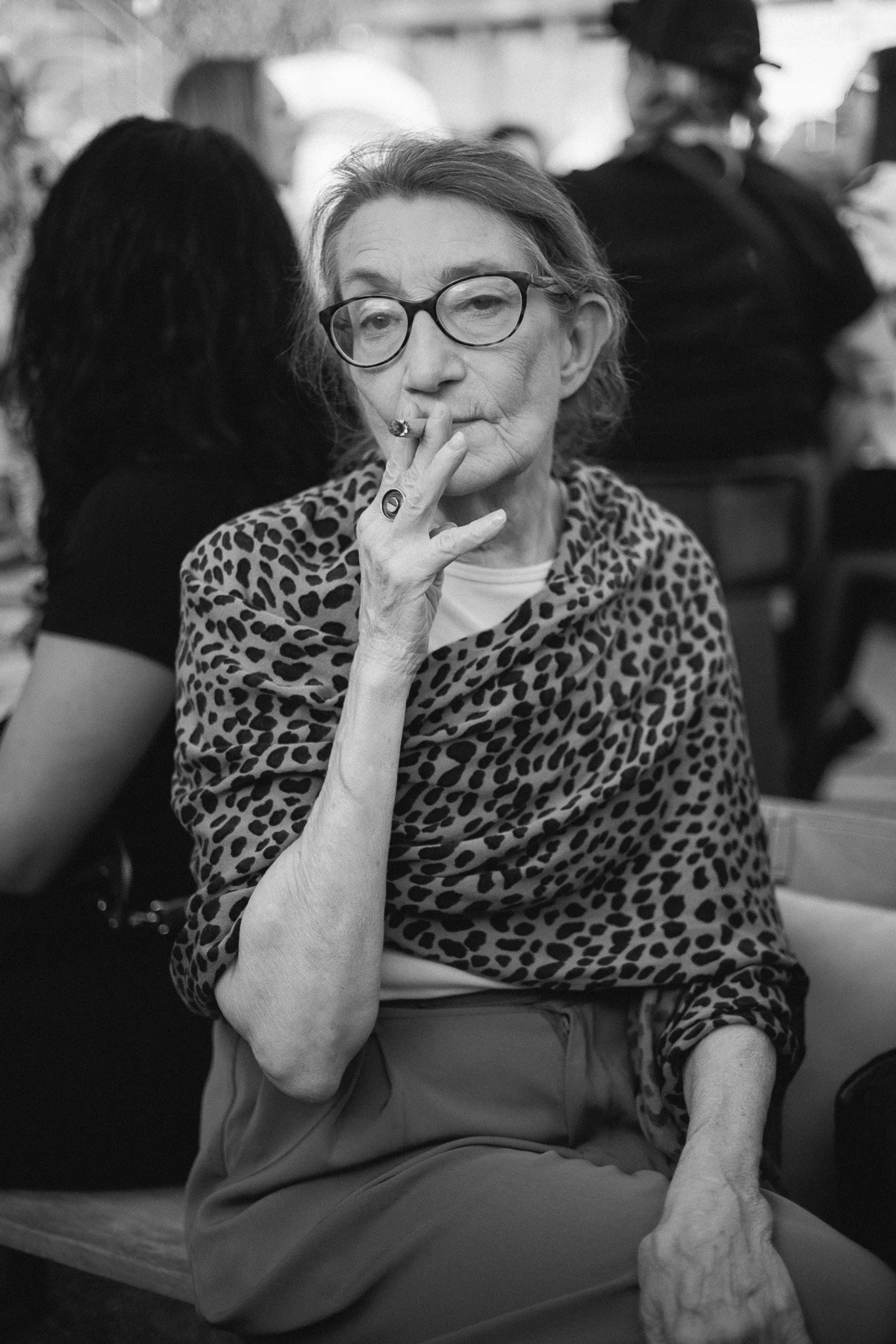An elderly woman with glasses, smoking a cigarette, wearing a leopard-print shawl and sitting in a social setting.