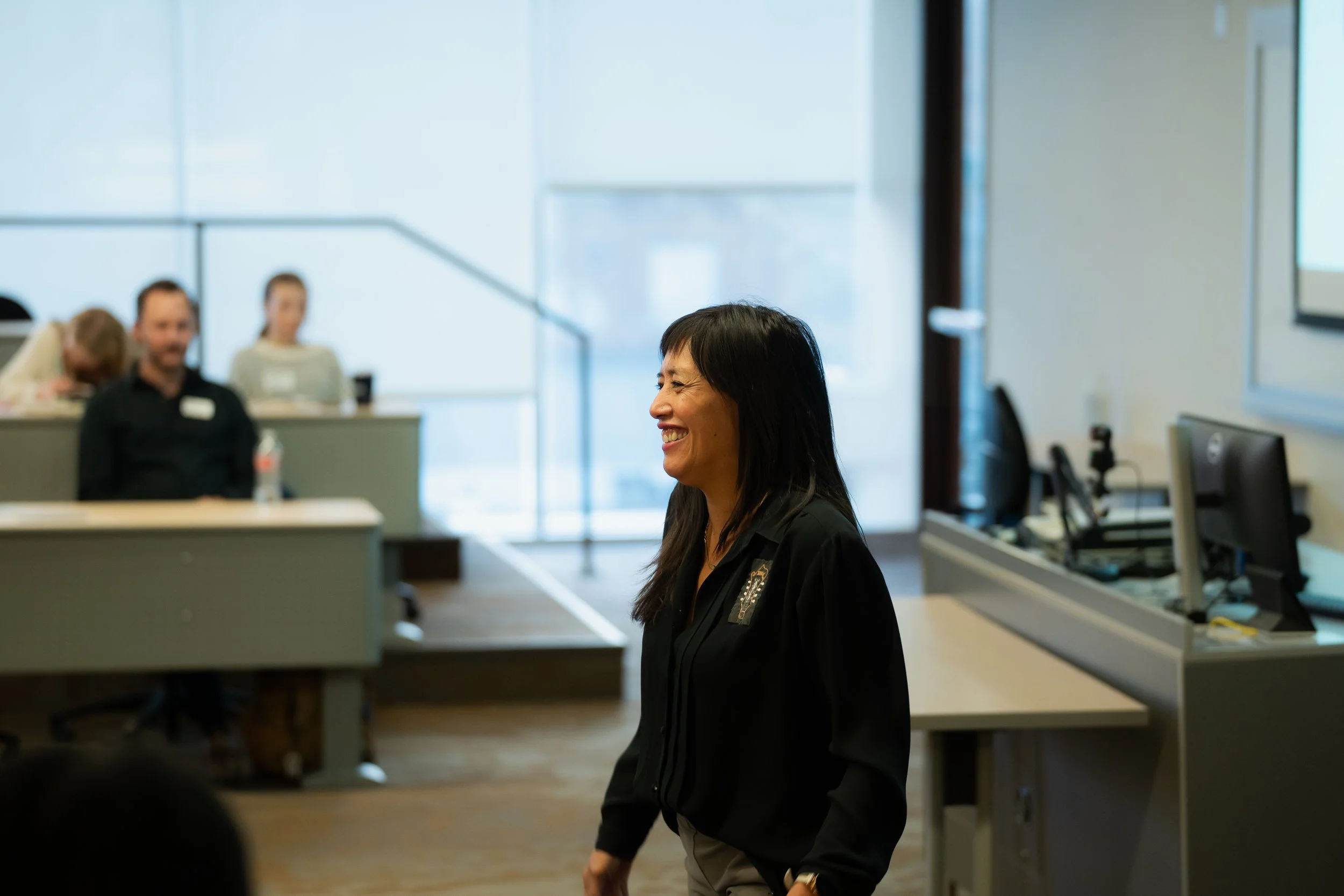 A woman smiling and standing in a conference room with other people seated in the background, wearing a black jacket, near a computer monitor and presentation screen.