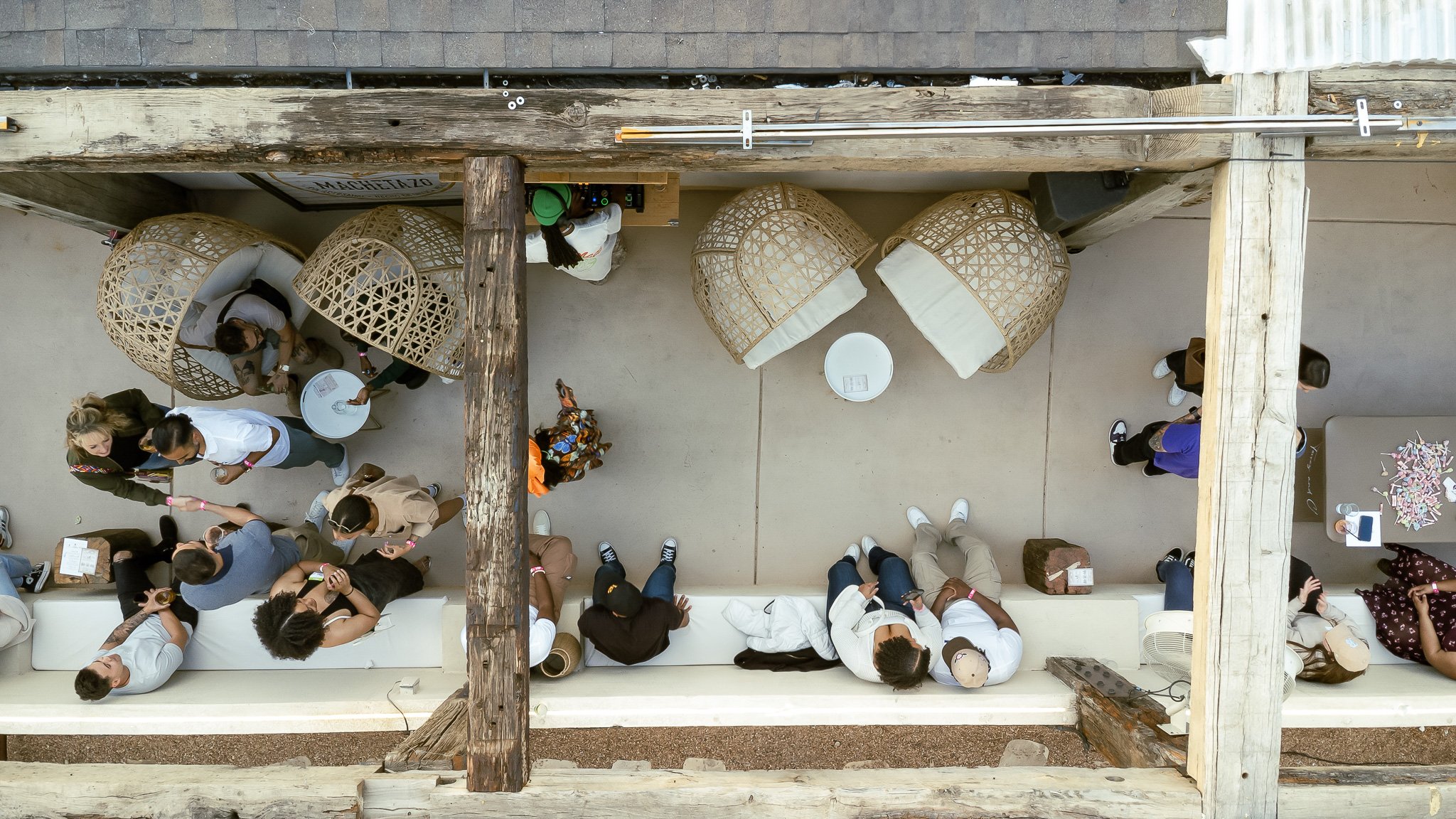 An aerial view of a rooftop gathering with people chatting, some sitting on a long white couch, others standing, surrounded by wicker bubble chairs and a table with a pile of torn paper, against a background of wooden beams and part of the building's