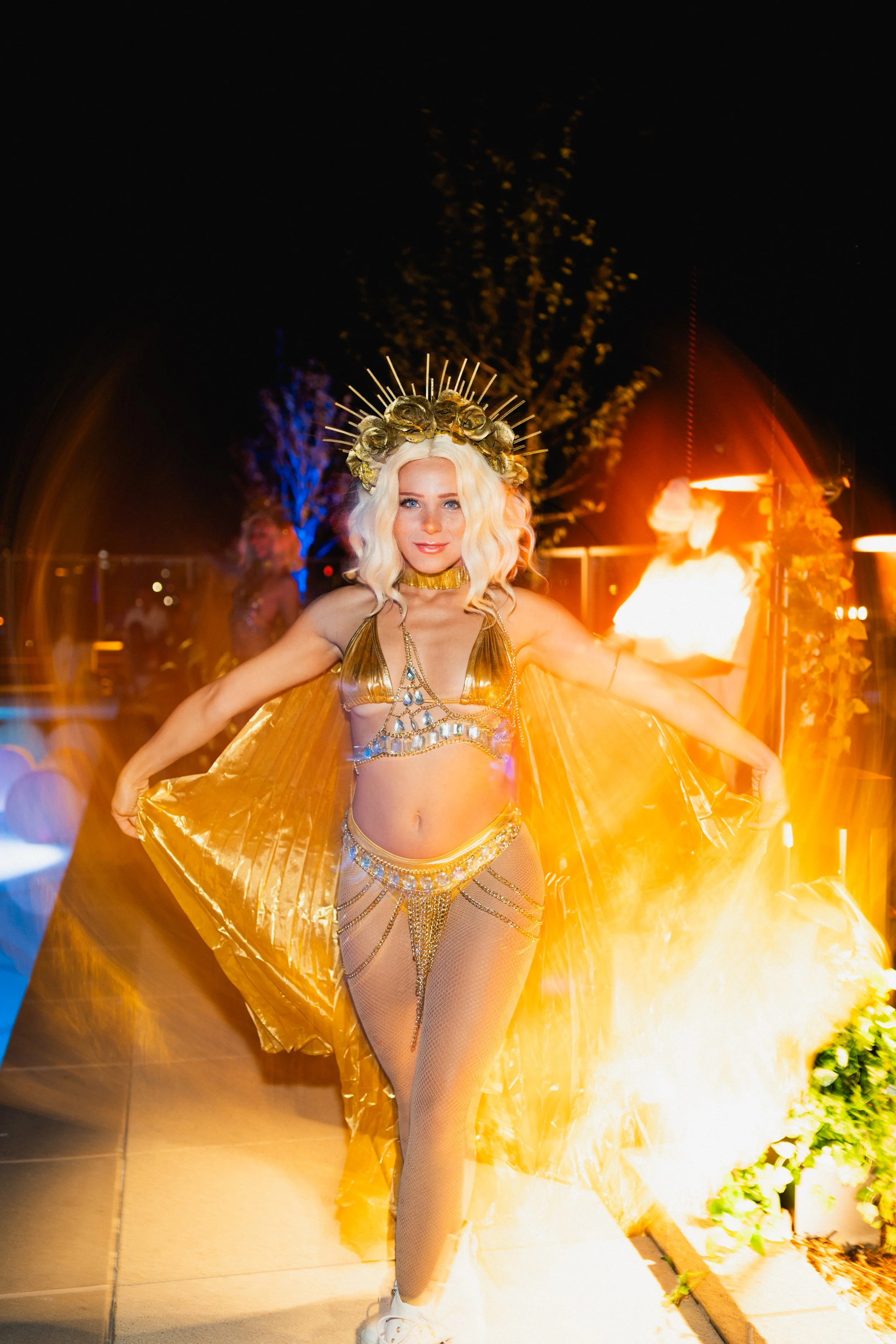 Woman dressed in a gold outfit with a crown, standing in front of a fire display at night.