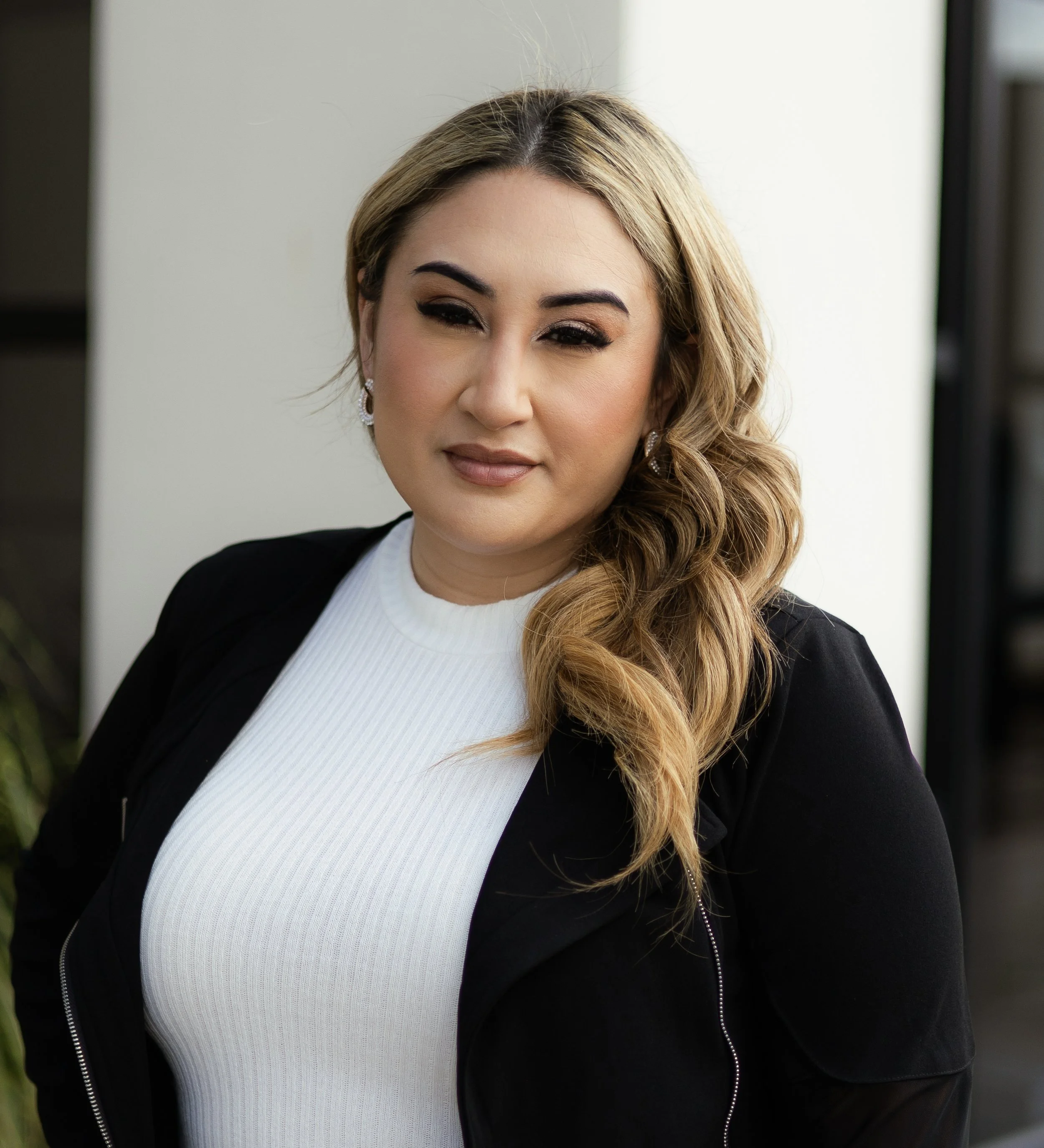 A woman with blonde, wavy hair wearing a white top and black jacket, standing indoors with a neutral background.