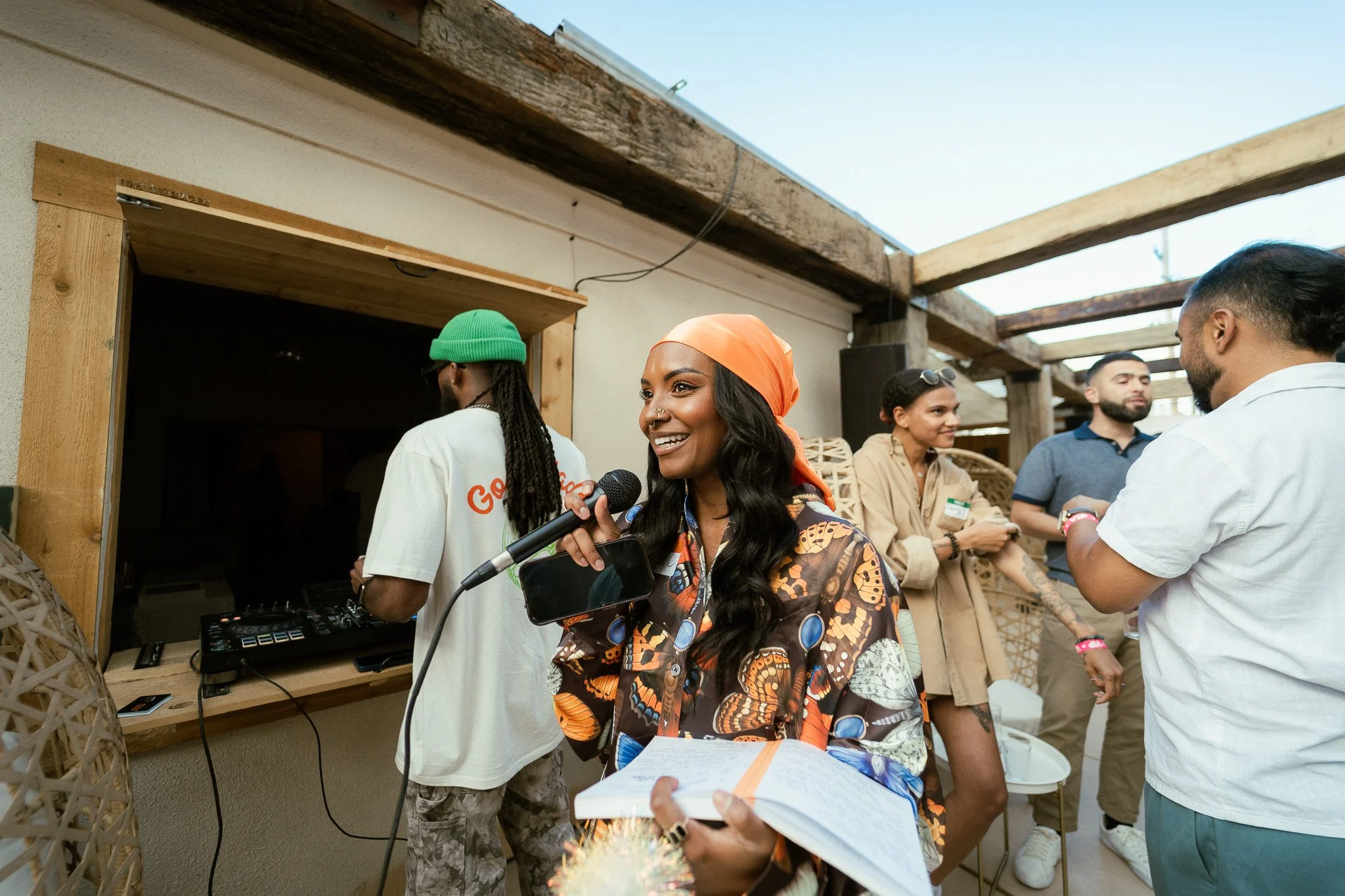 People socializing outdoors under a partially open wooden structure, with a woman in the center holding a microphone, smiling, and surrounded by others chatting.