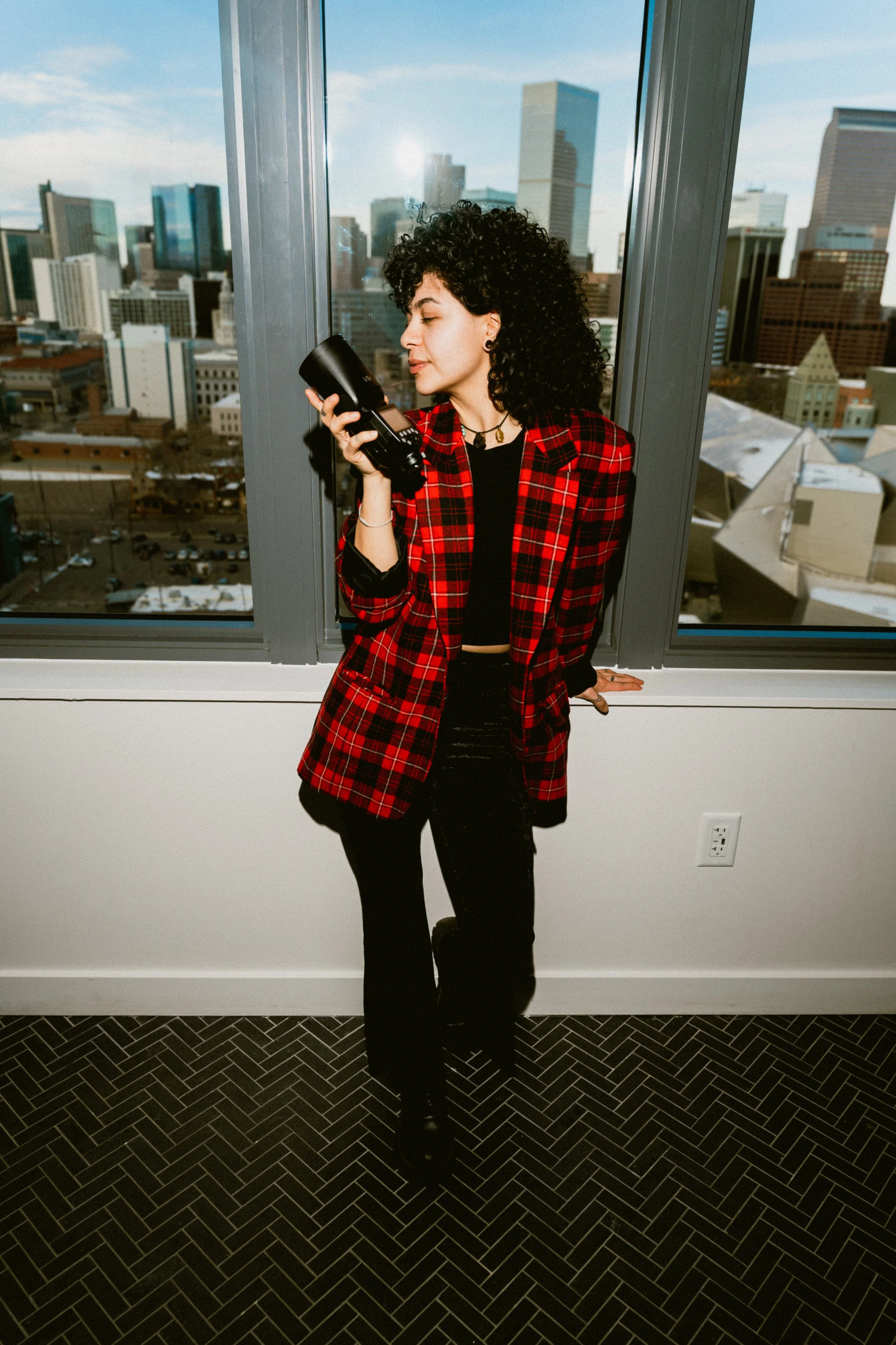 A woman with curly dark hair wearing a red plaid blazer and black pants, standing by a large window in a city building, holding a camera and looking at it with her eyes closed.