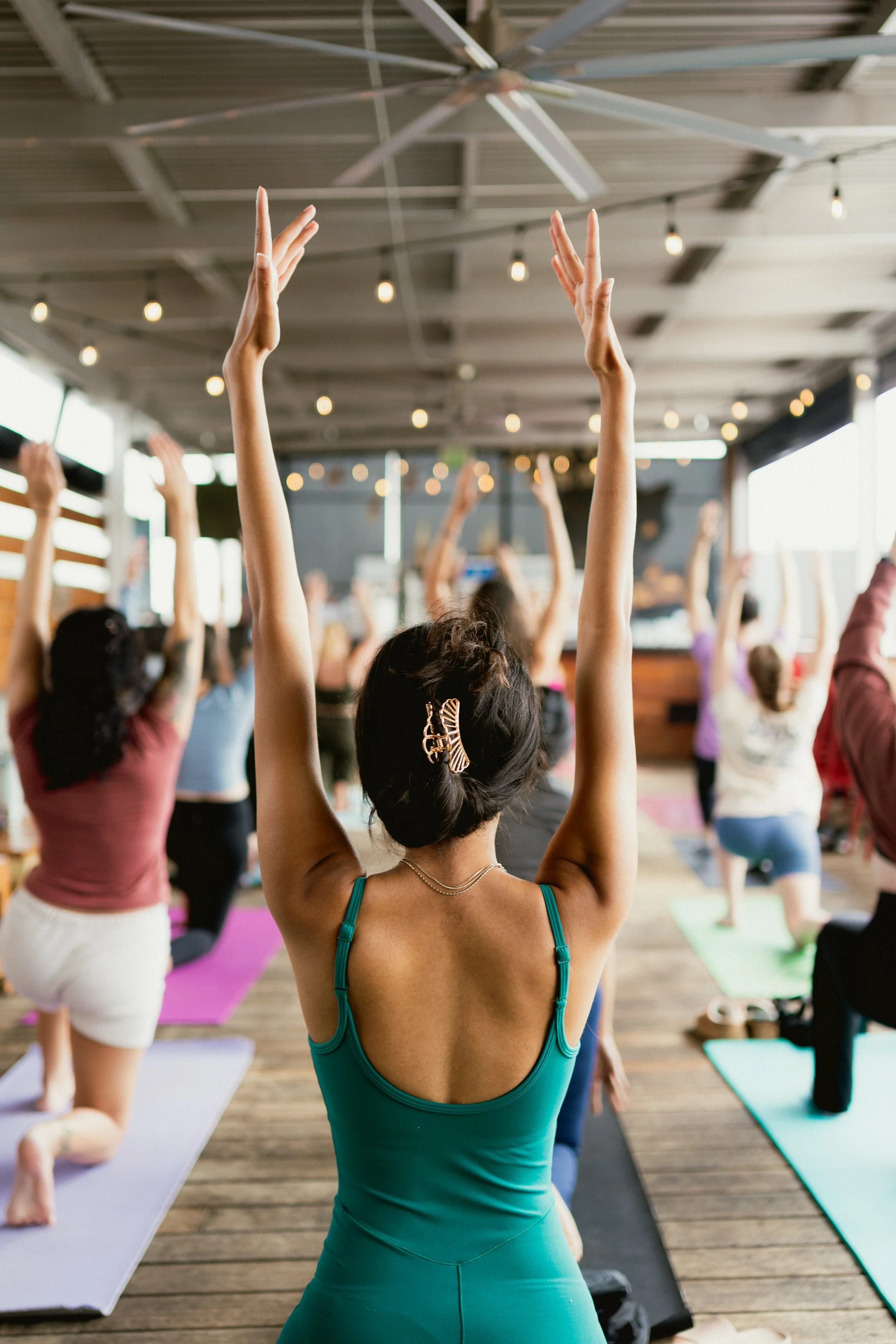 Group of people participating in a yoga class on yoga mats, led by instructor at the front, in a semi-outdoor setting with string lights hanging from the ceiling.