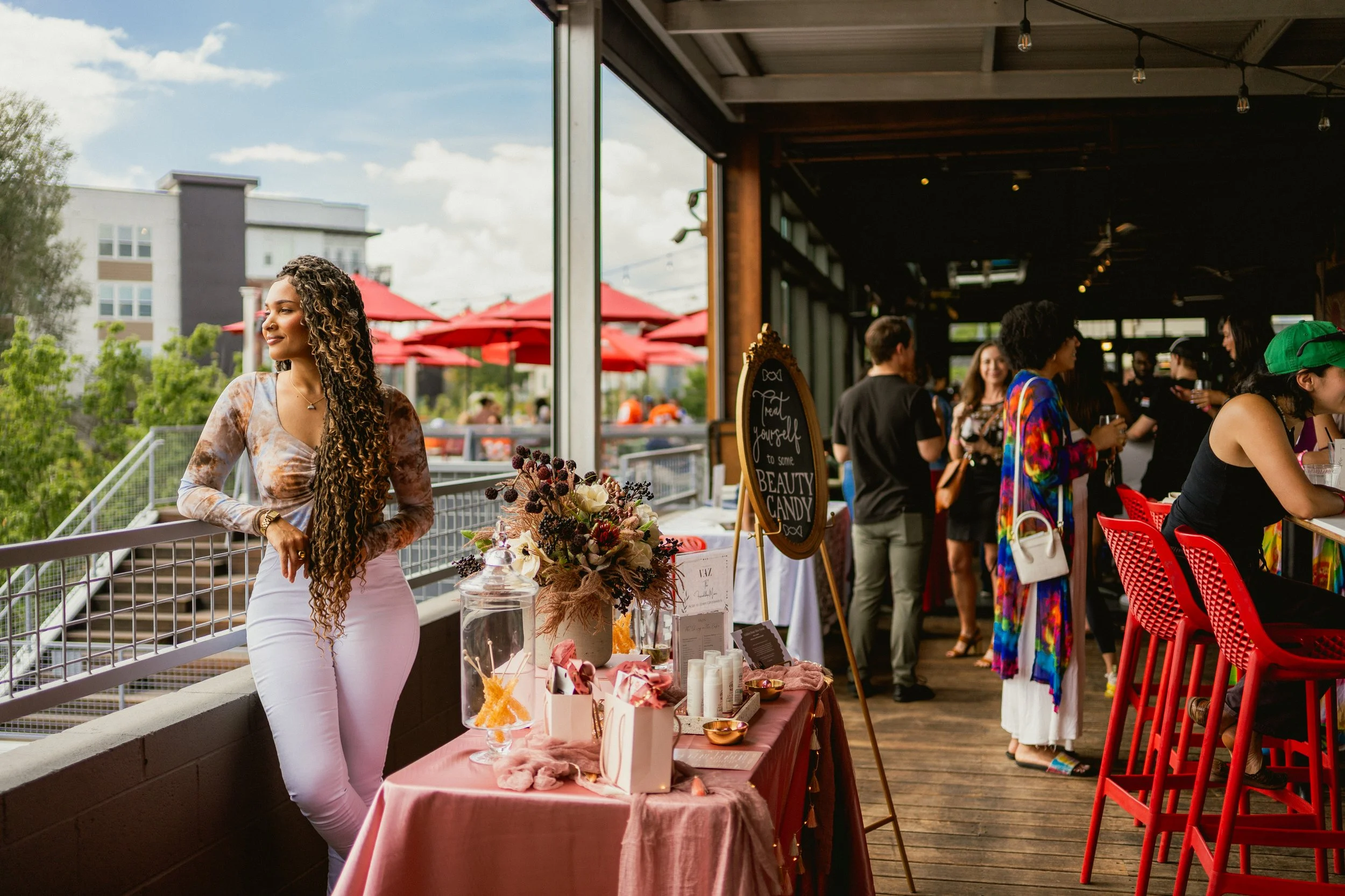 A woman with long curly hair standing near a booth with a sign that says 'Treat Yourself to Some Beauty Candy,' with a table displaying flowers and beauty products, in an outdoor-to-indoor event space with other people socializing in the background.
