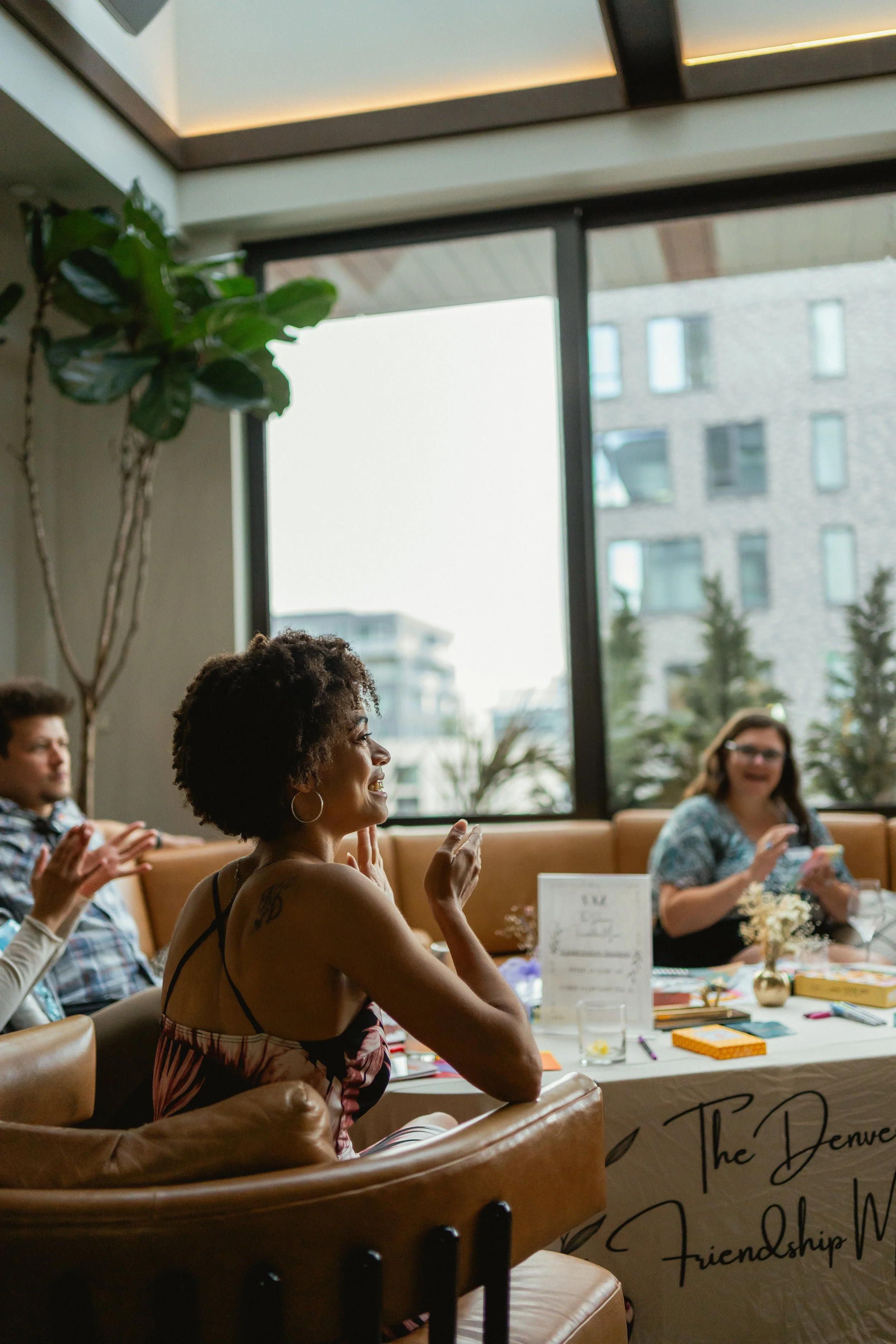 Group of women sitting at a table during a friendly gathering or event, smiling and engaging, indoor setting with large windows and plants.