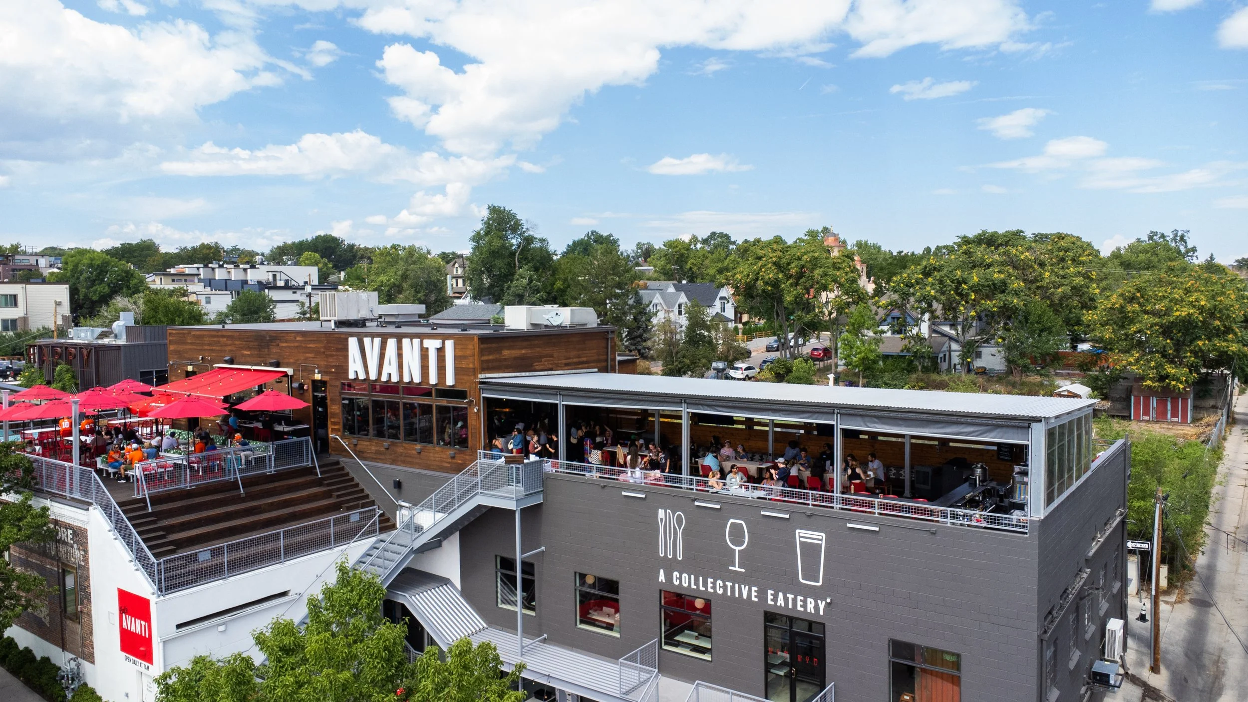 Aerial view of a multi-story restaurant with outdoor seating, including red umbrellas, and a sign that reads 'AVANTI.' The building has a modern design with large windows, and a grey facade with icons representing food and drink, labeled 'A COLLECTIVE EATERY.' There are trees and residential houses in the background under a blue sky with some clouds.
