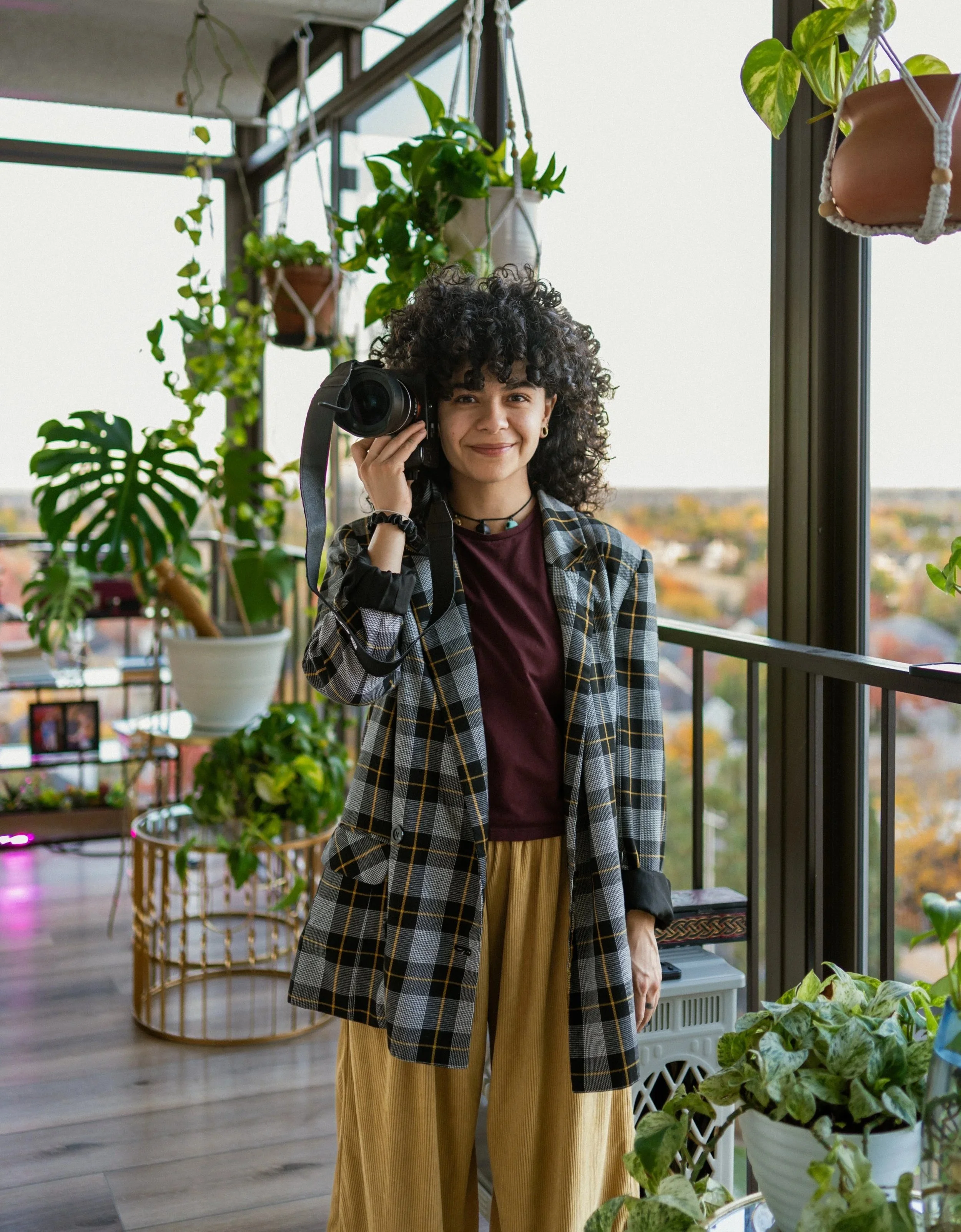 A woman with curly dark hair holding a camera near her face, standing in a room filled with hanging and potted plants next to large windows overlooking a cityscape during daytime.