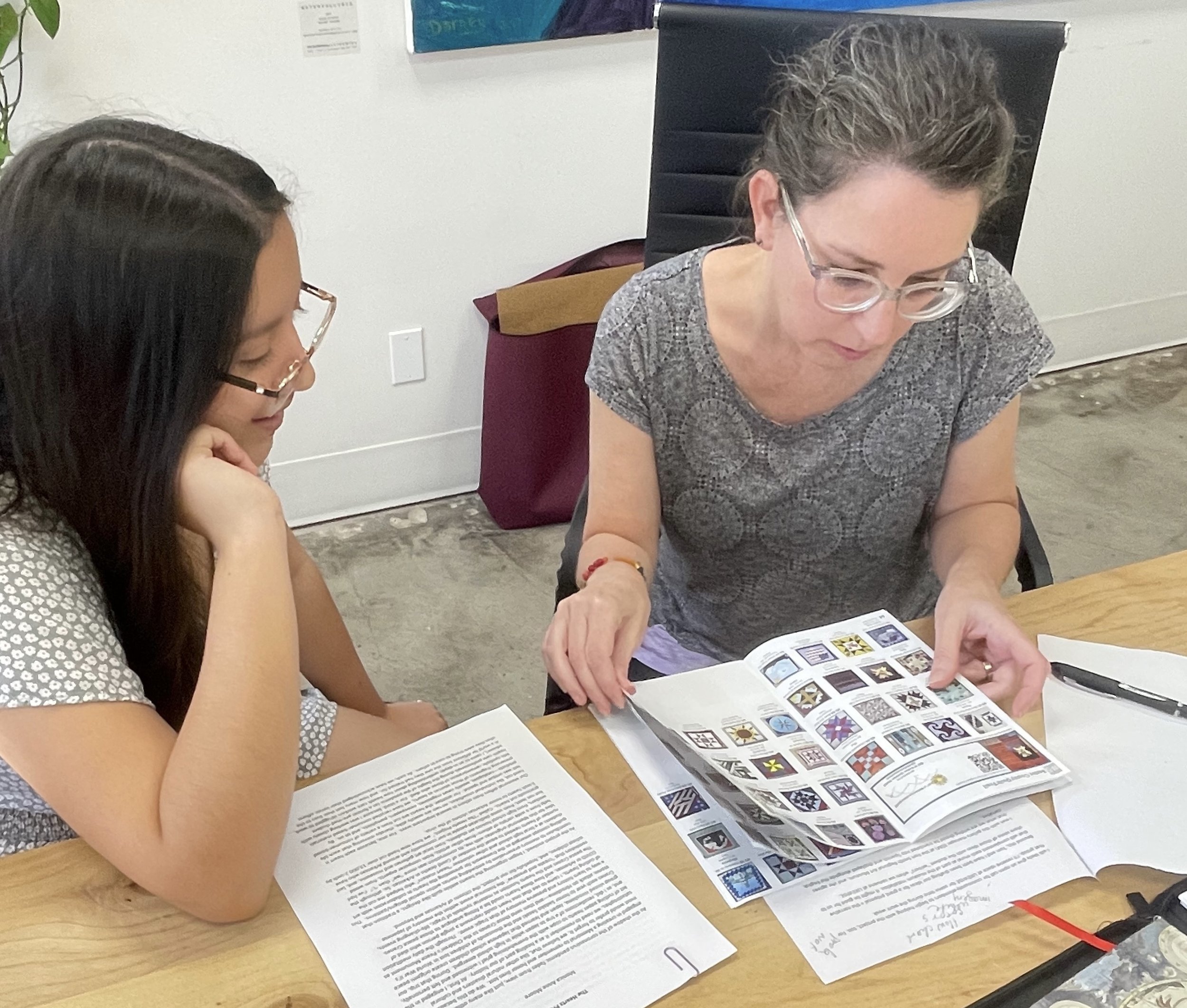 Monica Moore and Laura Avila sit together and study quilt making patterns in a book. Monica wears glasses and her brown and gray hair is swept back in a bun. Laura also wears glasses and has long, straight brown hair.
