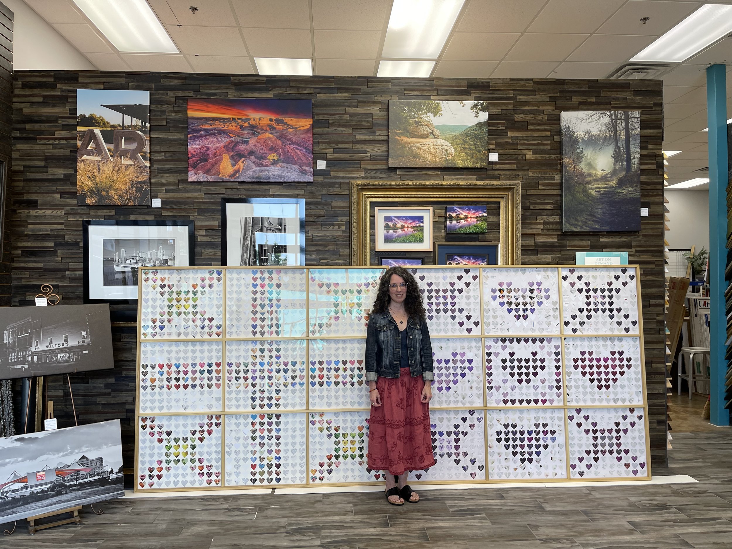 Monica Moore, artist, stands in front of the 6 foot panels of cut-out hearts, part of the Covid Hearts Project, in a framing shop. She is a white woman with long curly brown hair, a jean jacket and long flowy red skirt. Behind the large panels of her