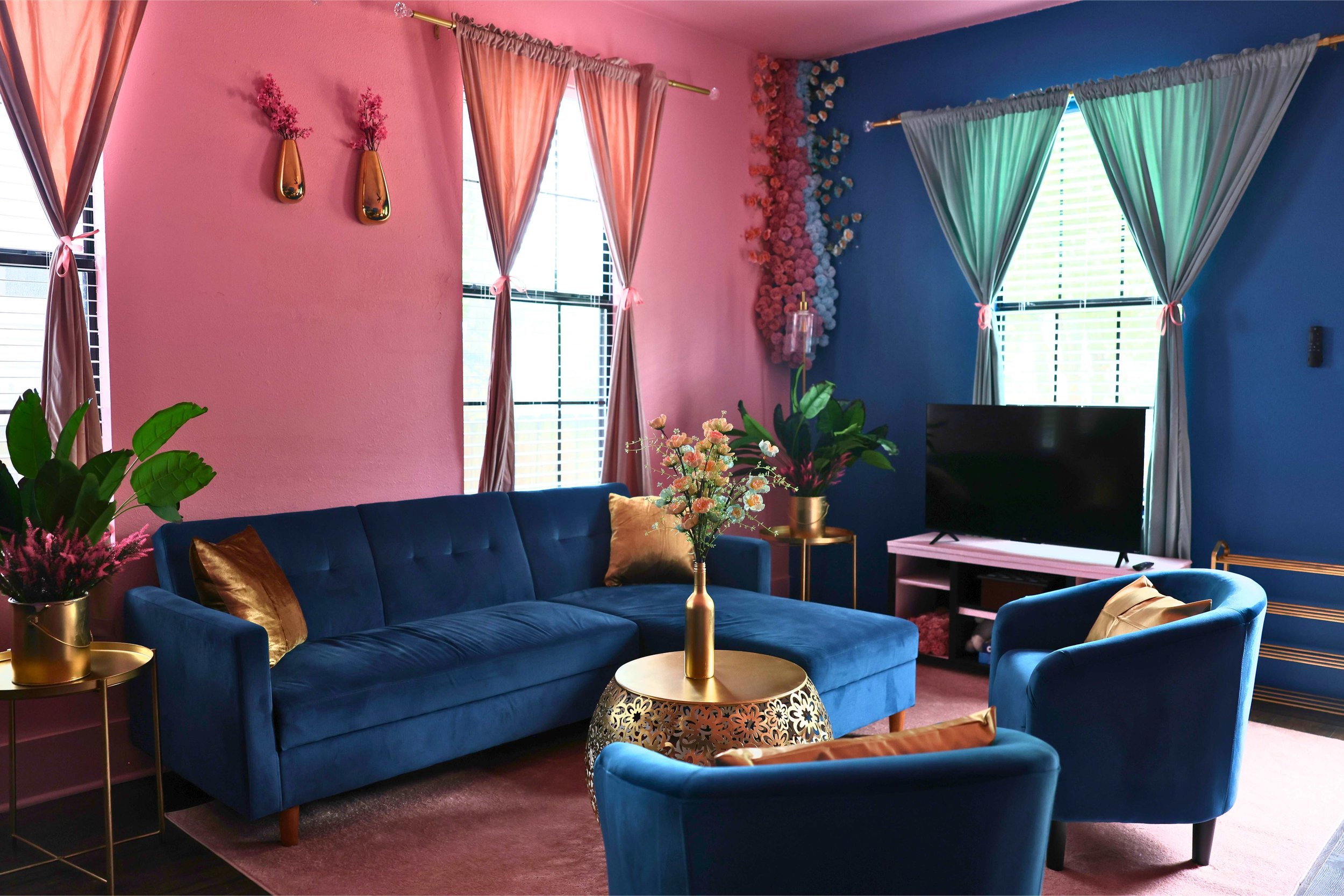 Colorful living room with blue and pink walls, featuring a blue couch and armchairs, gold accents, floral decor, and a TV.