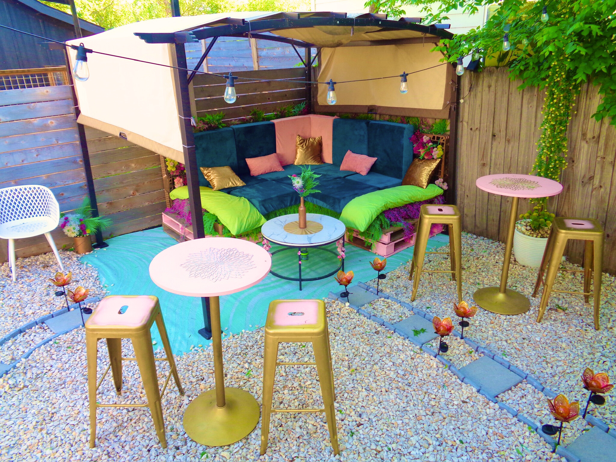 Outdoor patio setup with a canopy bed, colorful cushions, and small tables surrounded by decorative lighting and plants.