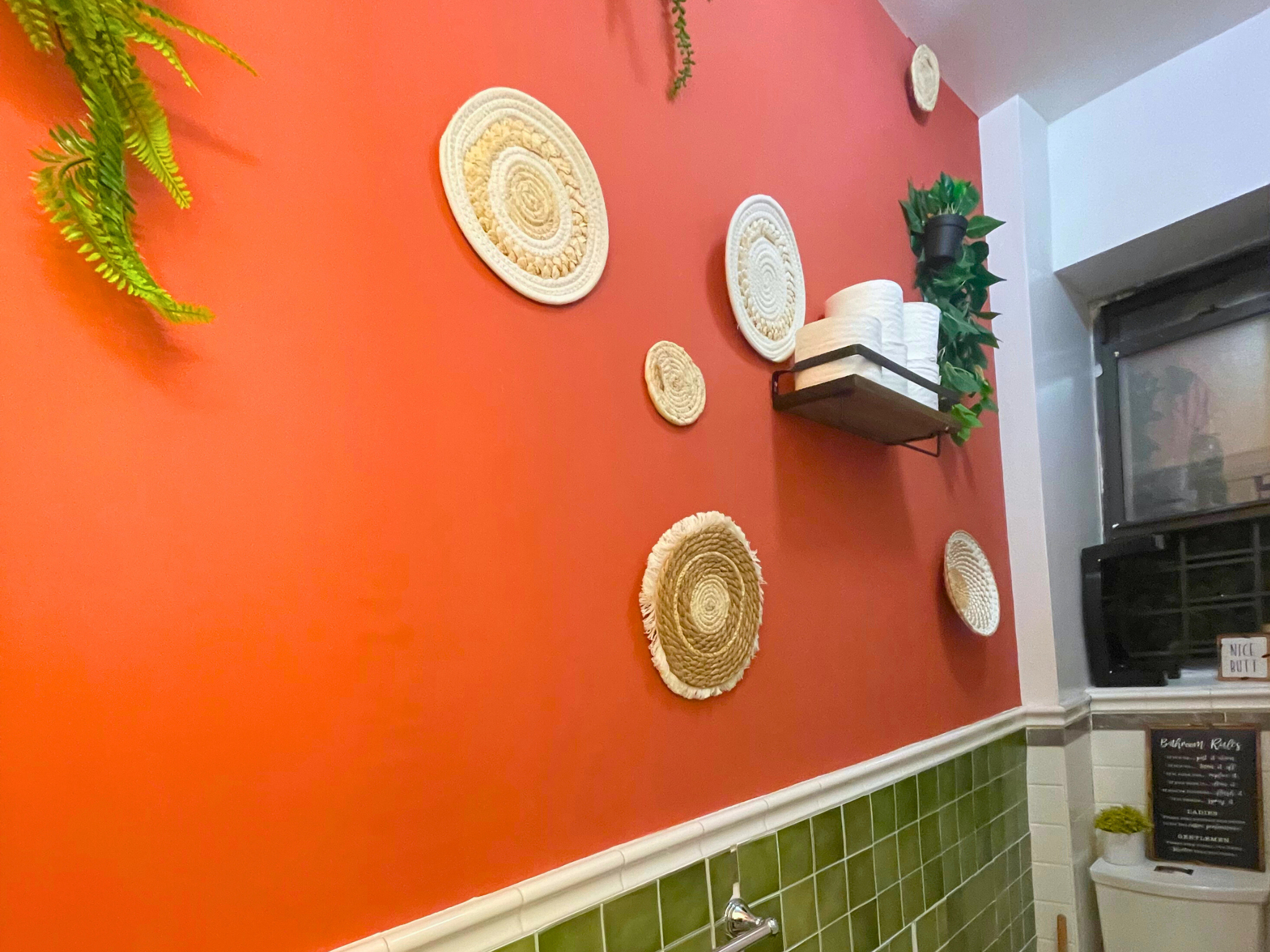 Bathroom with red wall adorned with woven circular decorations, a shelf holding white rolls, greenery, and a window with "Nice Butt" sign. Below, green tiles and a small blackboard with bathroom rules.