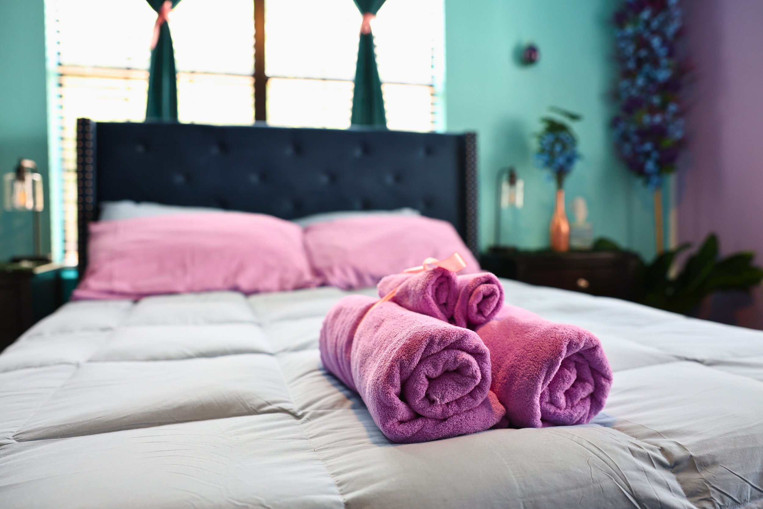 Bedroom with a neatly made bed featuring a dark upholstered headboard and pink pillows. Folded pink towels are placed on the bed, tied with a ribbon. The room has turquoise walls and decorative elements, including a vase with flowers.