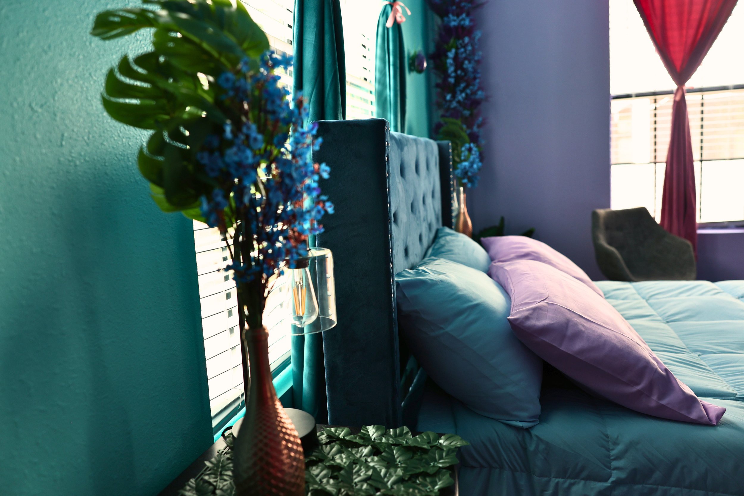 Colorful bedroom with blue and purple bedding, teal walls, and decorative flowers on a side table. A large window with red curtains and a green chair are visible.