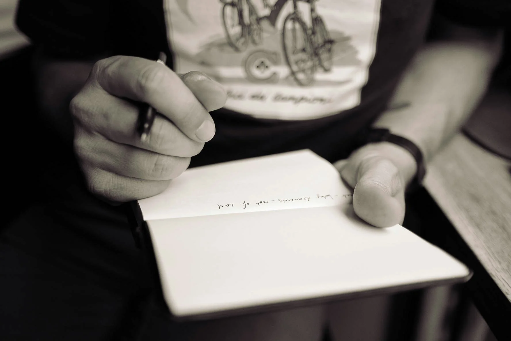 Close-up of a person writing in a notebook with a pen, focusing on their hand and a part of their T-shirt with bicycle illustrations.