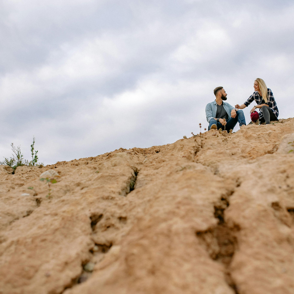 A man and woman sitting on a rocky hill, sharing an intimate moment, with the woman offering the man a ring or small object.