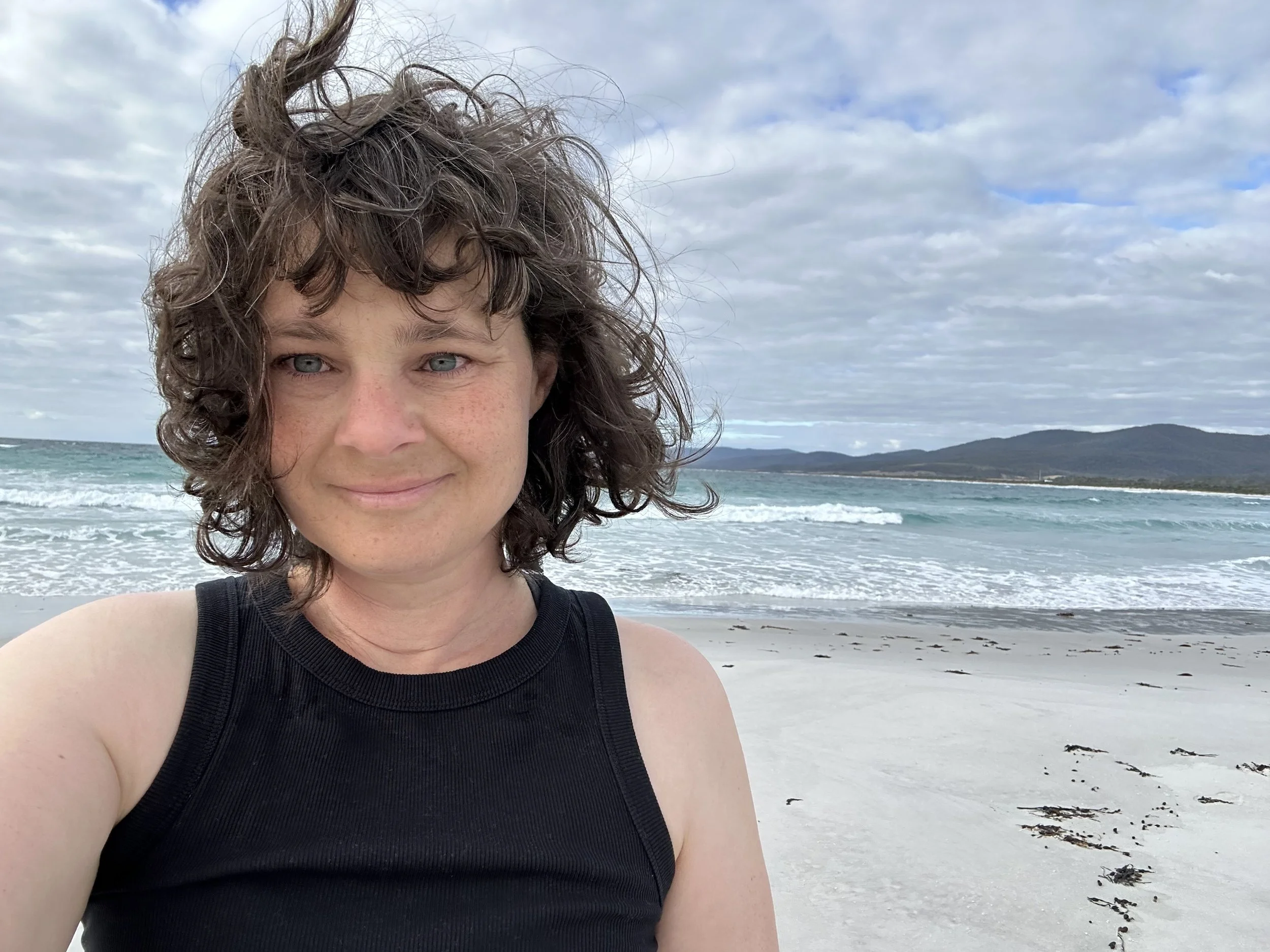 Woman with curly short hair smiling at the camera on a beach, with the ocean and cloudy sky in the background.