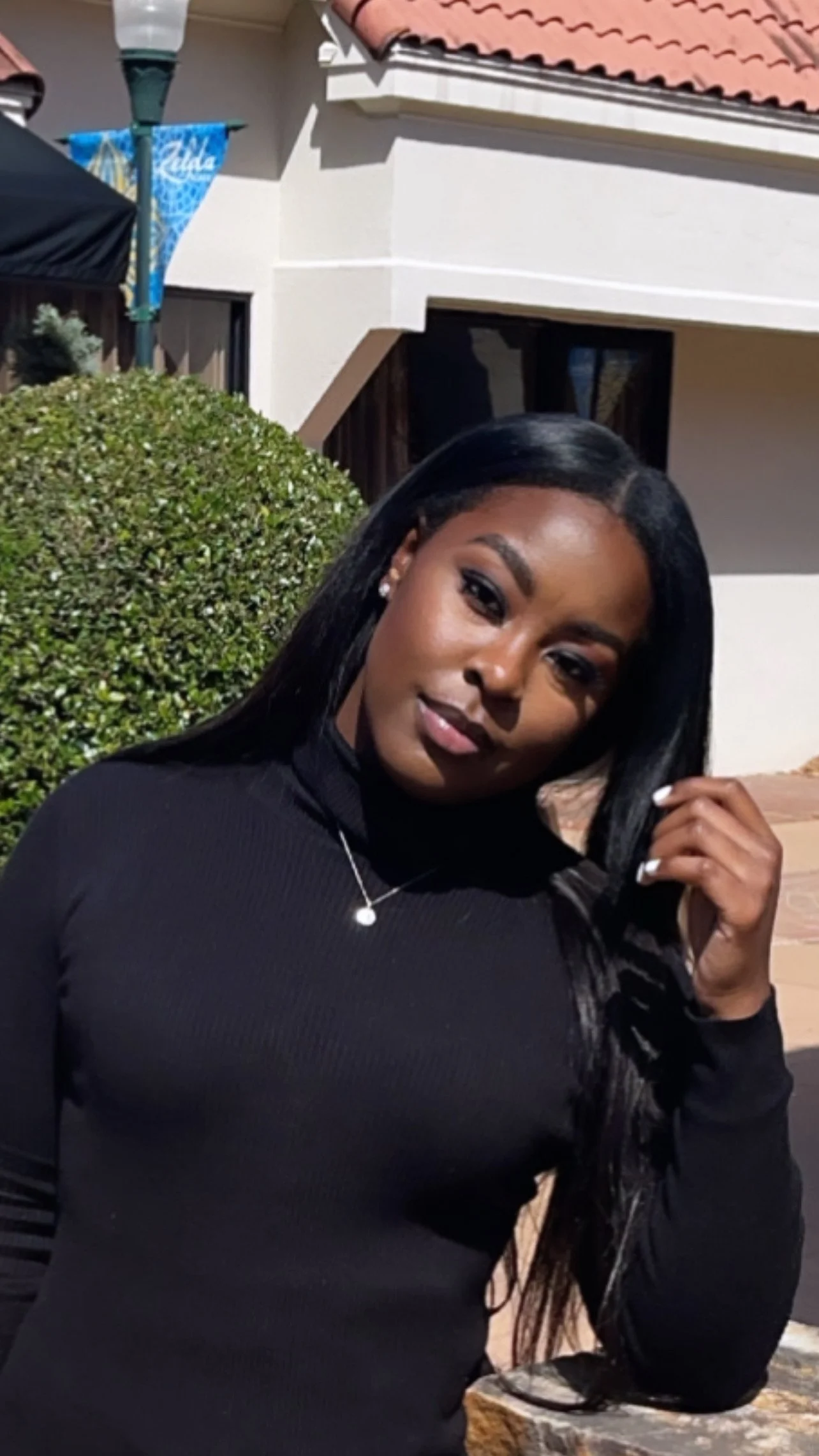 Person with long black hair, wearing a black top, standing outside near a building with a red-tiled roof and greenery.
