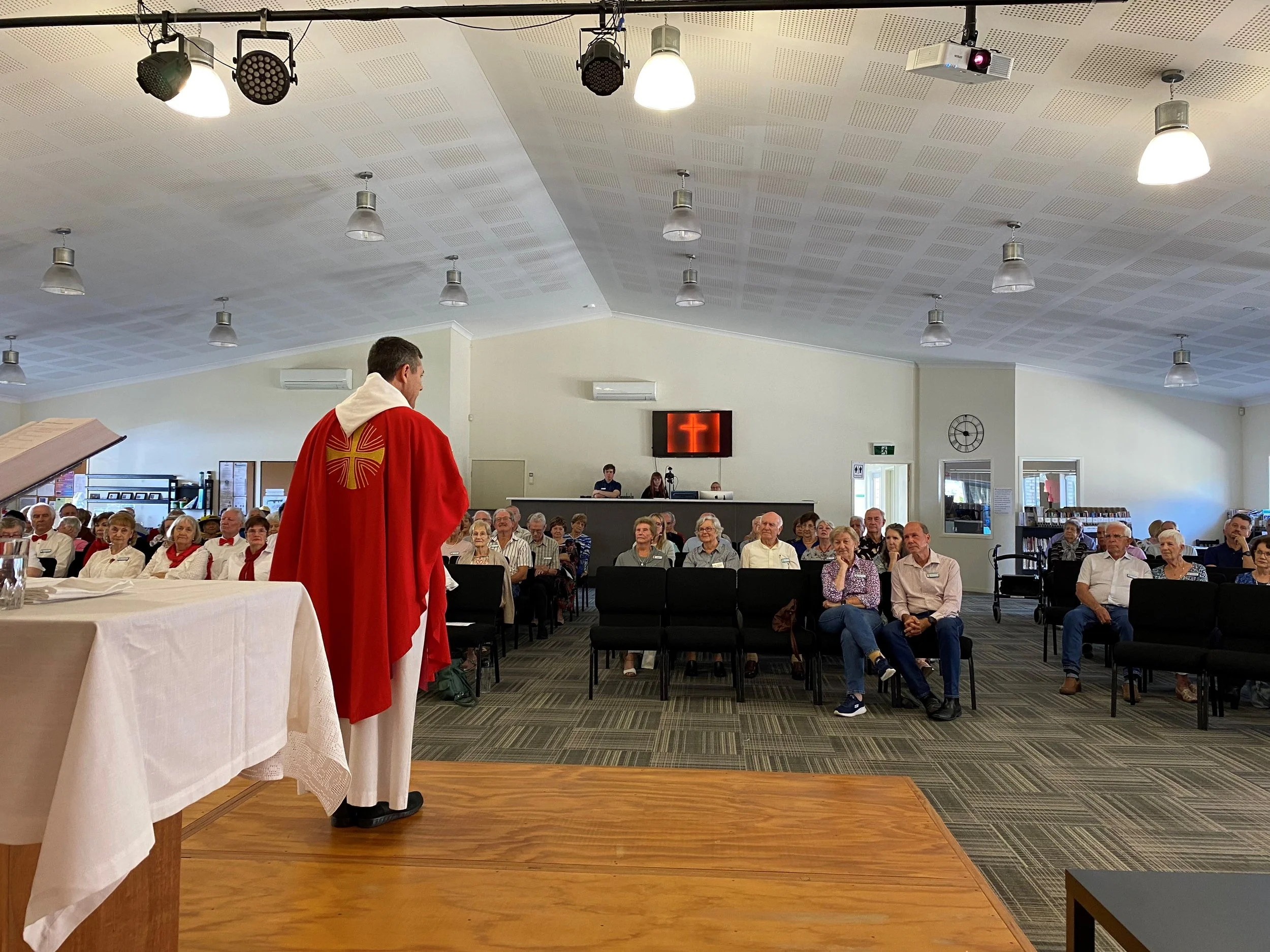 A priest or clergy member in red robes stands in front of a seated congregation in a church or community hall. The congregation of mostly elderly people watches attentively. There is a table with a white cloth and open book or book stand on the left.