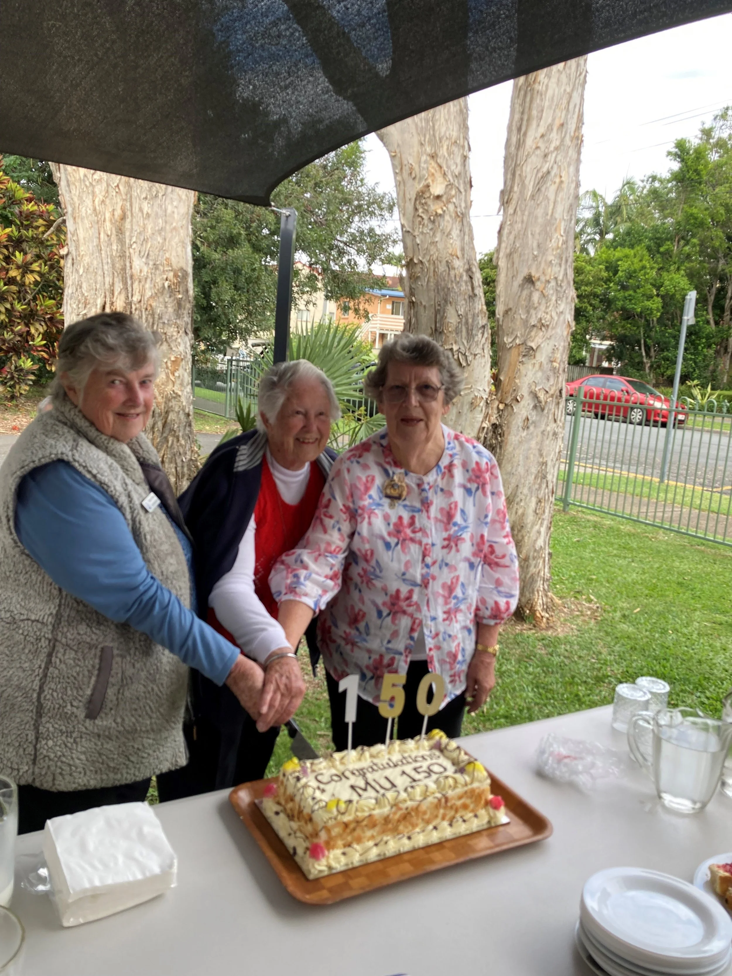 Three women celebrating a 150th anniversary with a decorated cake that says 'Congratulations Miu 150' under a dark canopy, with trees, a street, and a red car in the background.