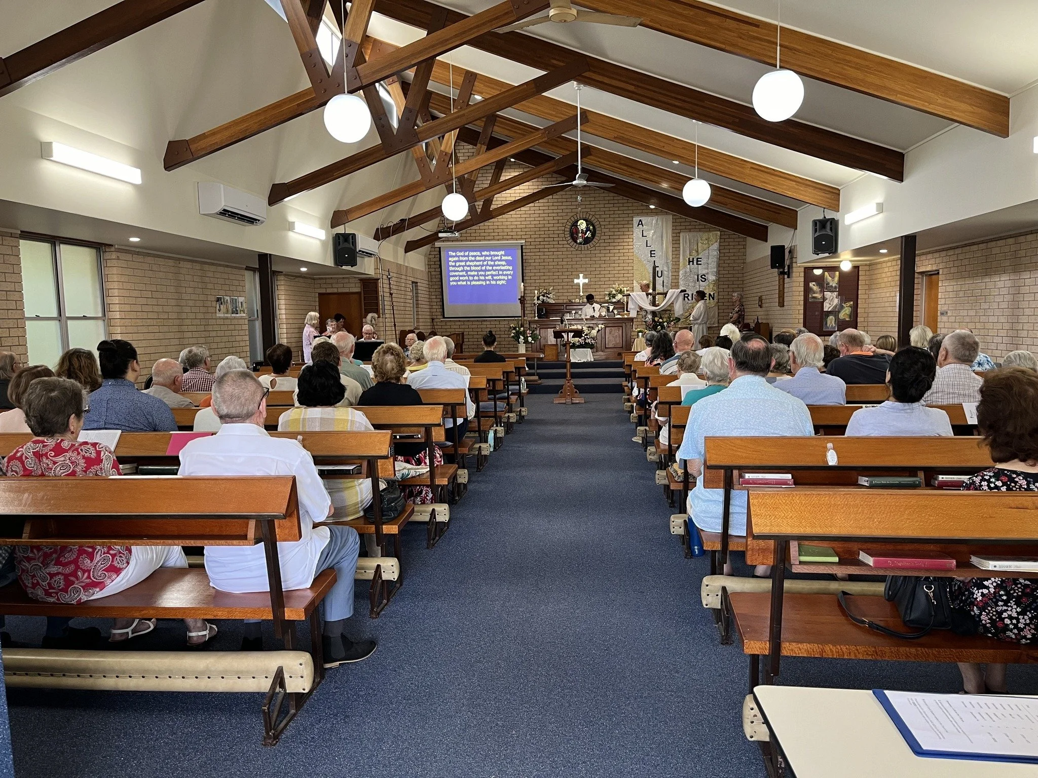 A congregation gathered inside a church with wooden beams on the ceiling, listening to a service at the altar. There are people sitting on wooden pews, and a presentation is displayed on a screen at the front.