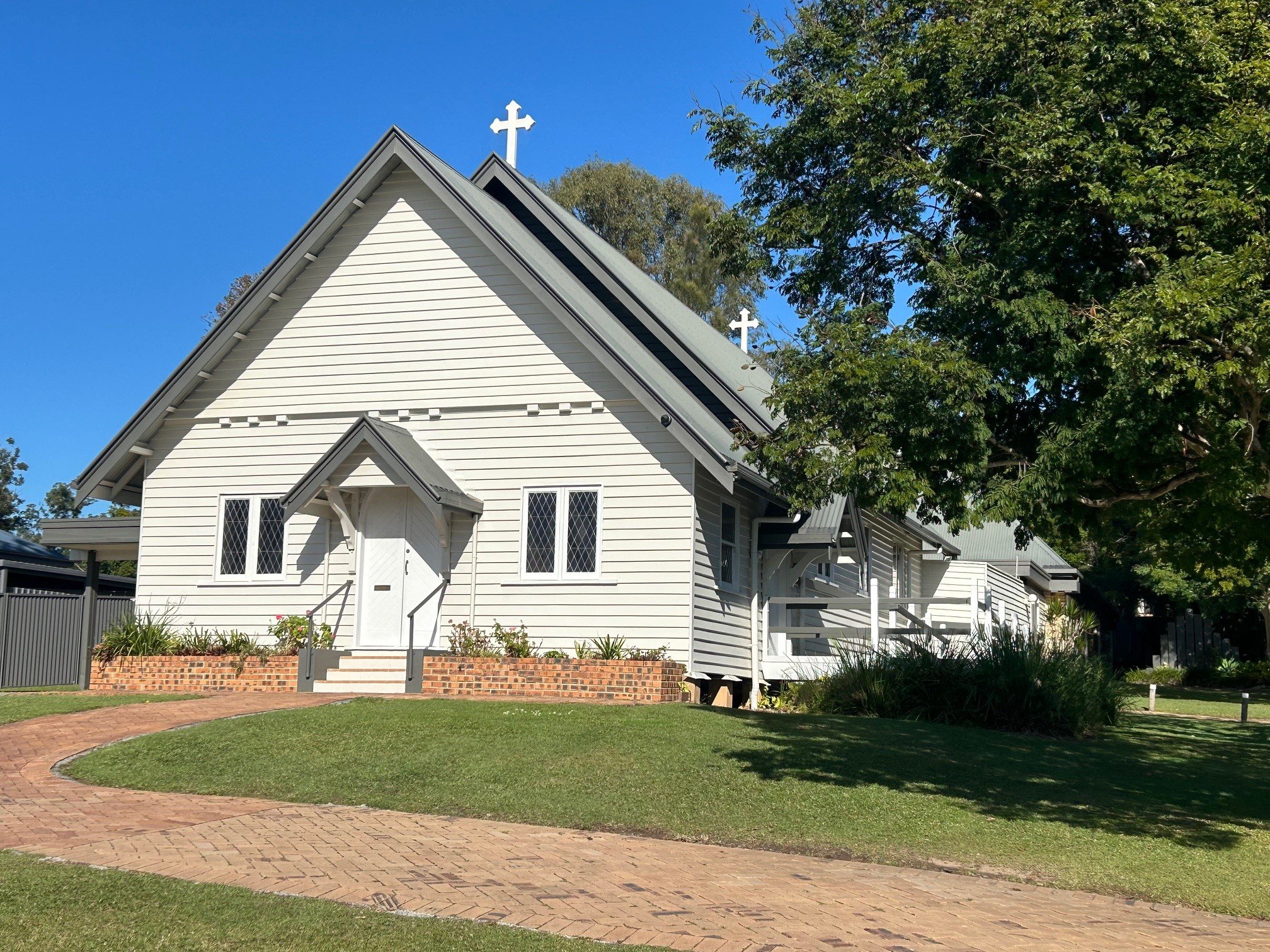 A white church with a steep roof, crosses on top, surrounded by trees, with a brick walkway leading to the entrance.