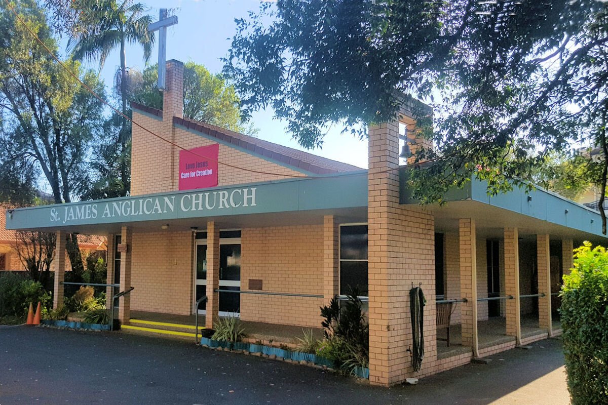 Front view of St. James Anglican Church, a brick building with a green awning, trees around, a cross on a pole, and a sign on the building facade indicating the church's name.