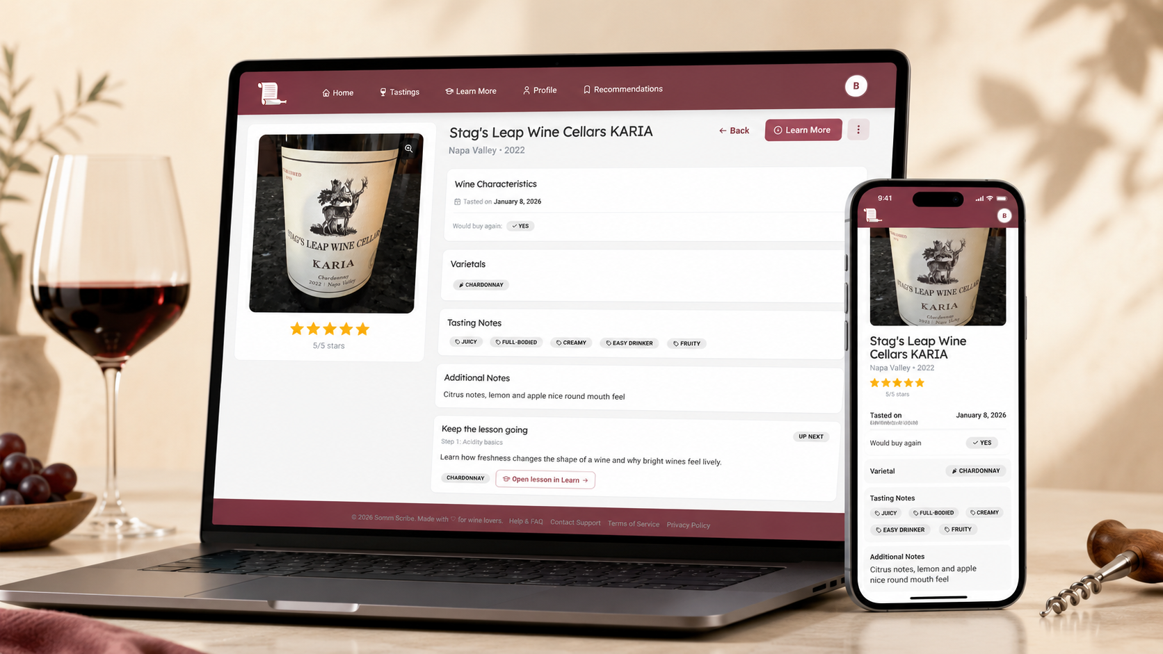 Laptop and smartphone displaying the Somm Scribe wine journal app on a wine tasting detail screen, set on a tabletop beside a glass of red wine, grapes, and a corkscrew.