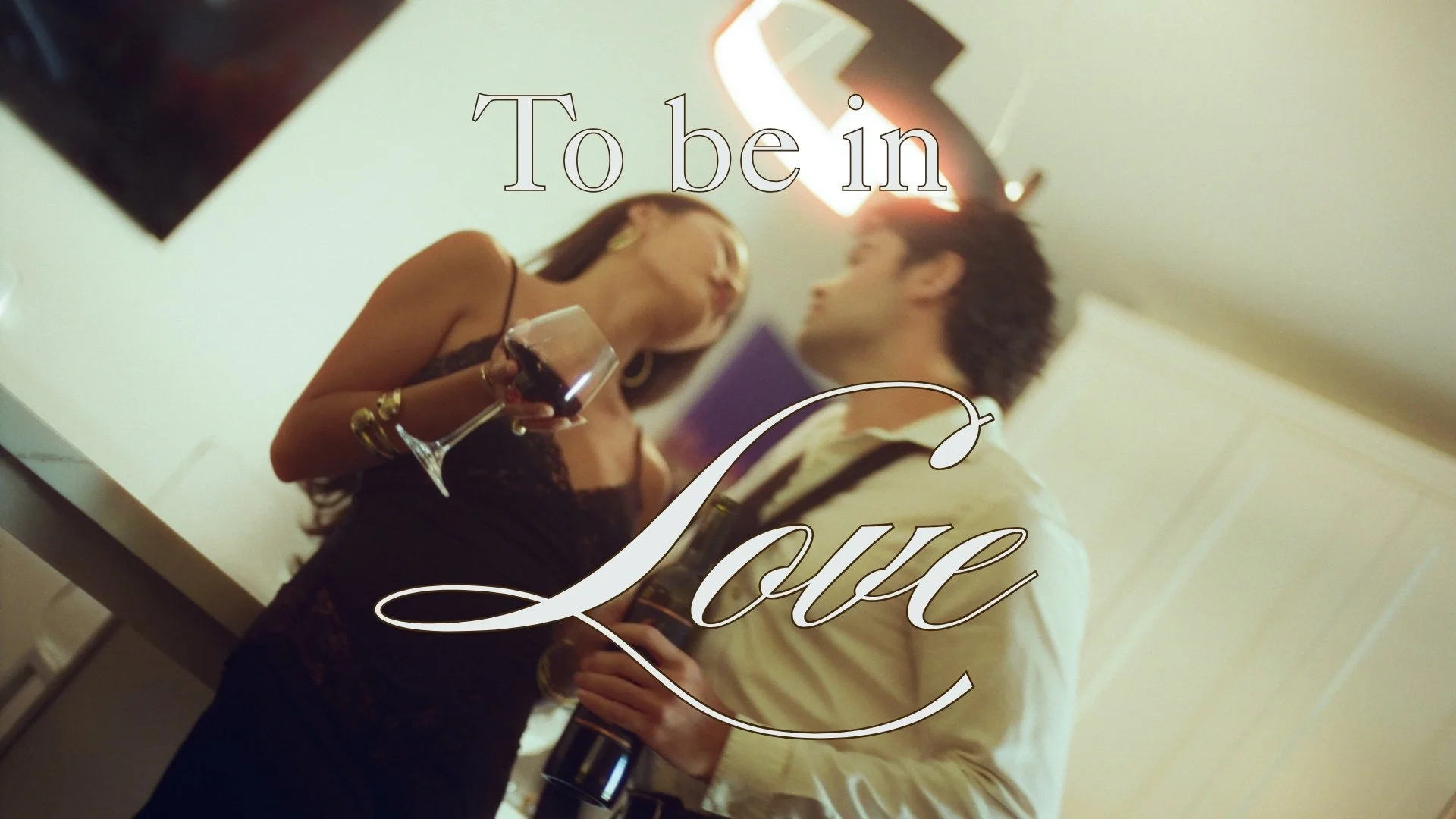 A woman holding a wine glass and a man holding a bottle of wine, standing close together at a social gathering, with the words 'To be in Love' overlaid on the image.
