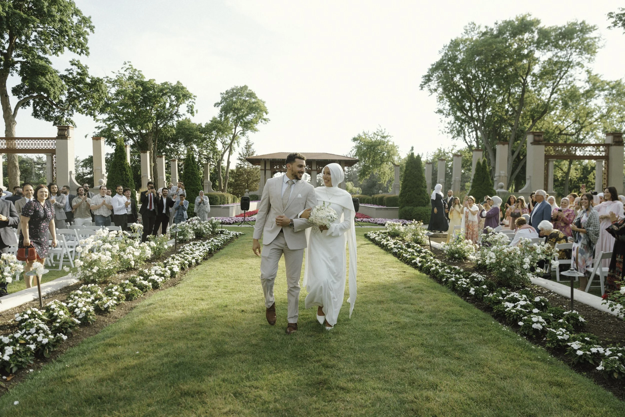 A bride and groom walking down an aisle outdoors during a wedding ceremony. The bride is wearing a white dress and hijab, holding a bouquet, and the groom is dressed in a beige suit. Guests are standing and seated, watching and smiling, with some taking photos. The setting is a lush, green garden with decorative pillars and trees.