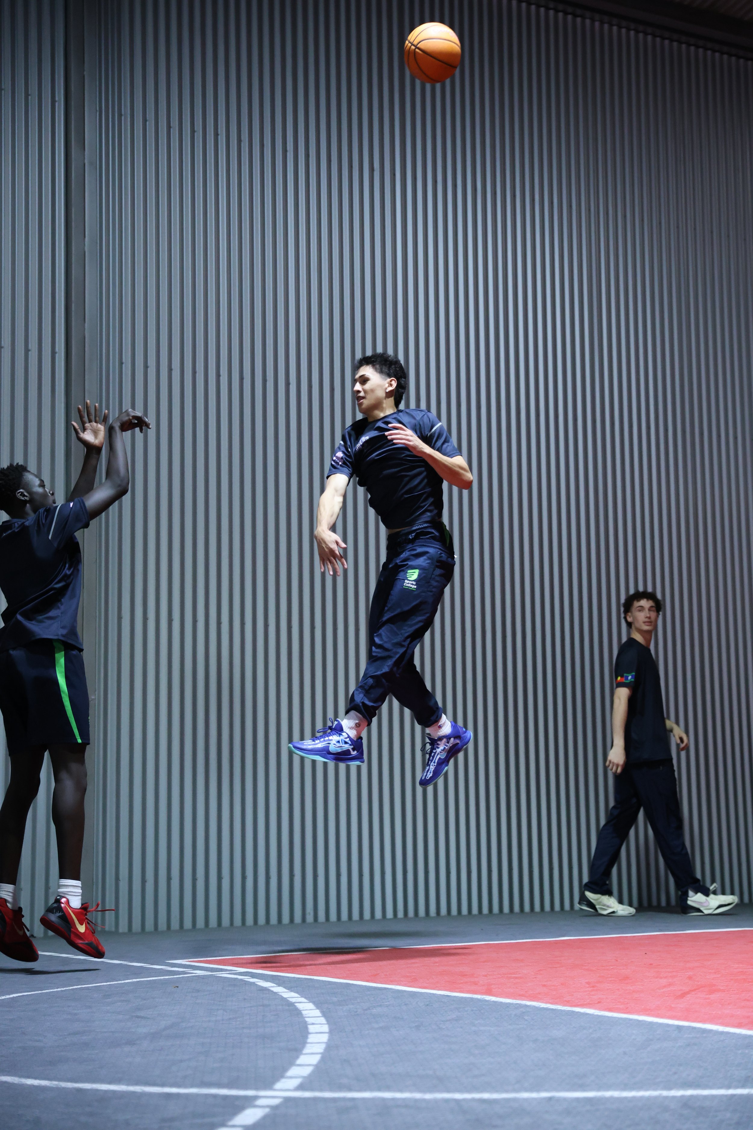 Two basketball players, one in mid-air jumping to make a shot, and the other reaching to block, on an indoor court with a metal wall background, with a third person standing to the side.