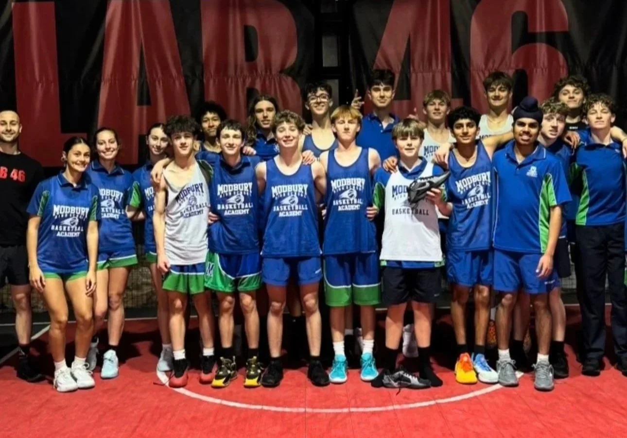 Group of young basketball players and coaches in team uniforms posing together for a photo on a basketball court, with some players holding sneakers.