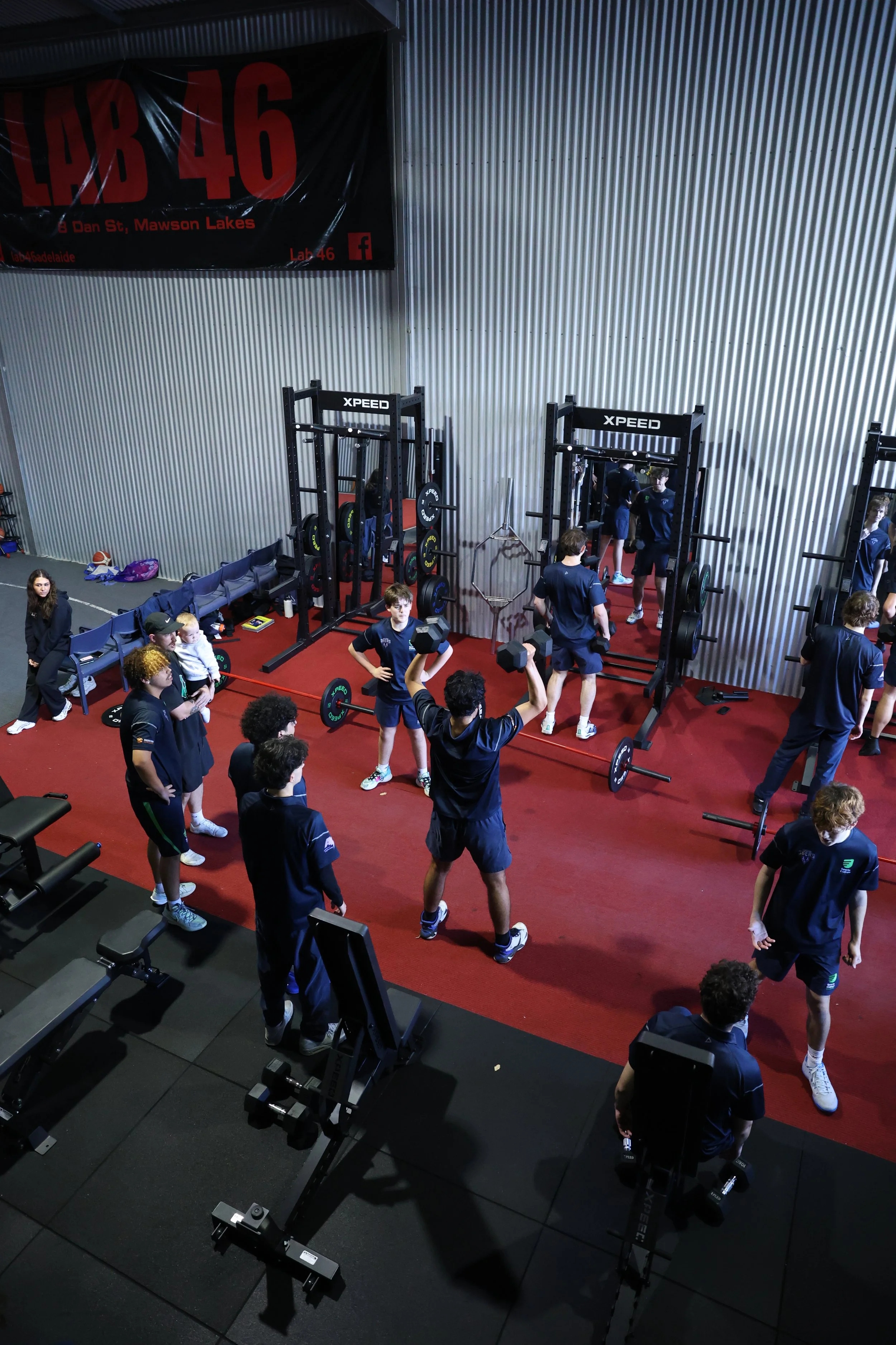 People working out and lifting weights in a gym with gray metal walls, black exercise equipment, and a red floor.